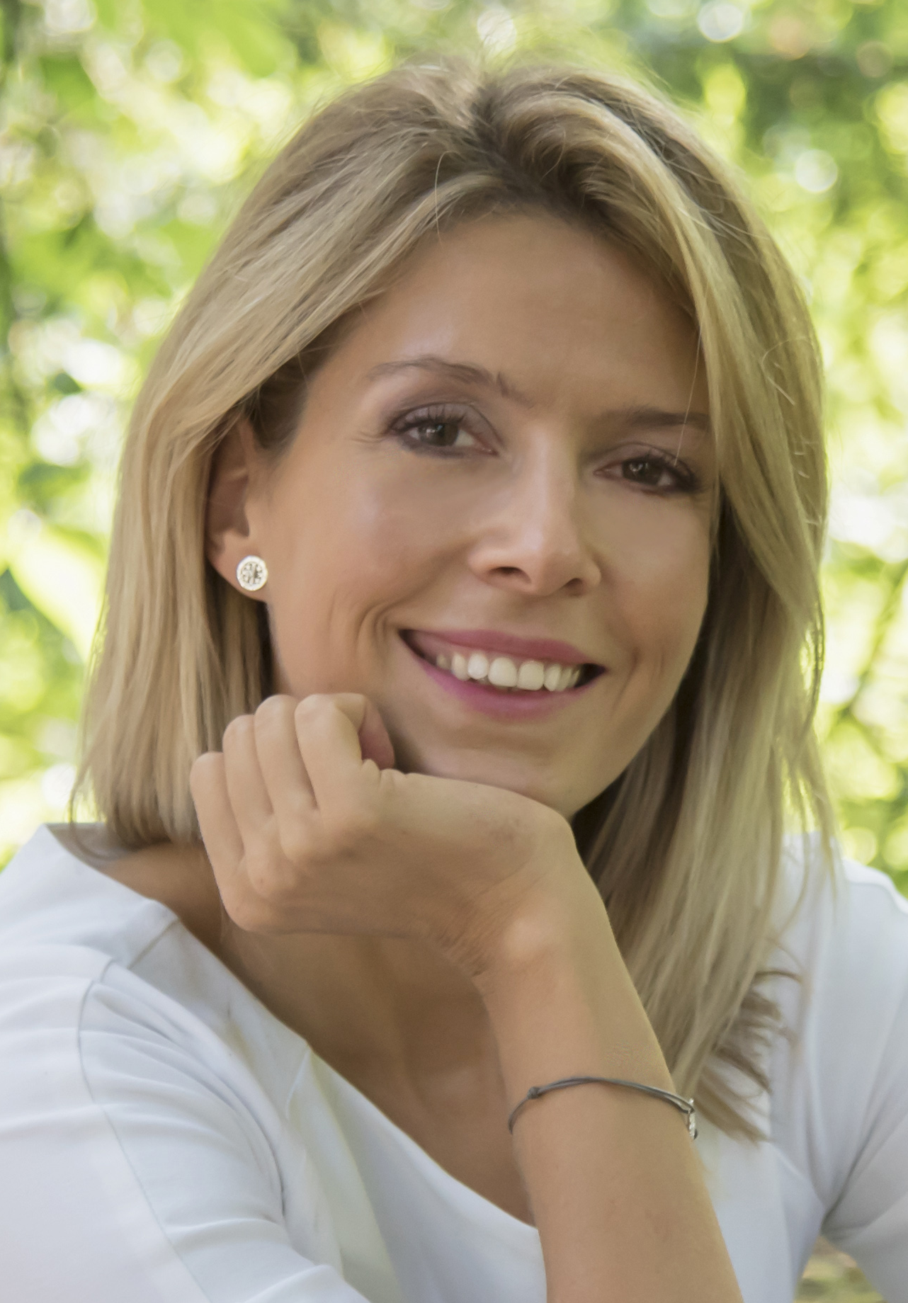 A woman with blonde hair smiling outdoors with greenery in the background.