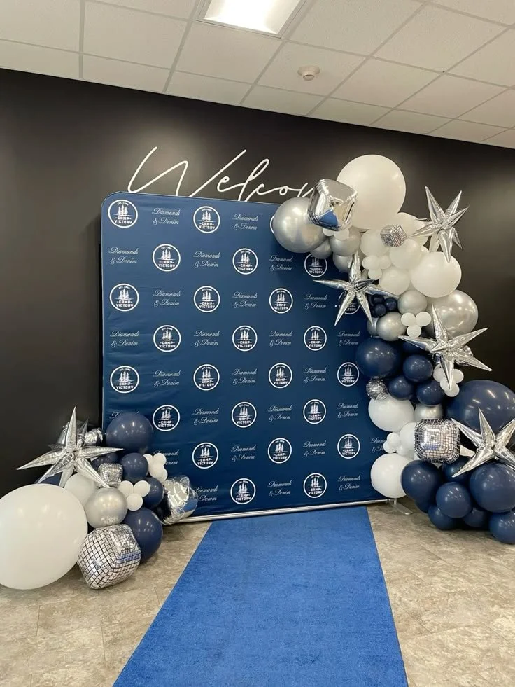 Balloon arrangement with white, navy, and silver balloons, including star-shaped and cube-shaped balloons, surrounding a blue backdrop with 'Diamond & Daven' logo and a partial 'Welcome' sign, leading to a blue carpet.