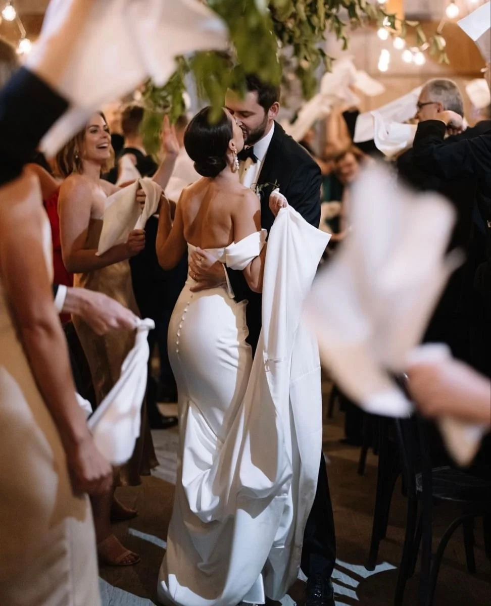A bride and groom share a kiss under a decorated arch while guests throw white paper napkins in celebration.
