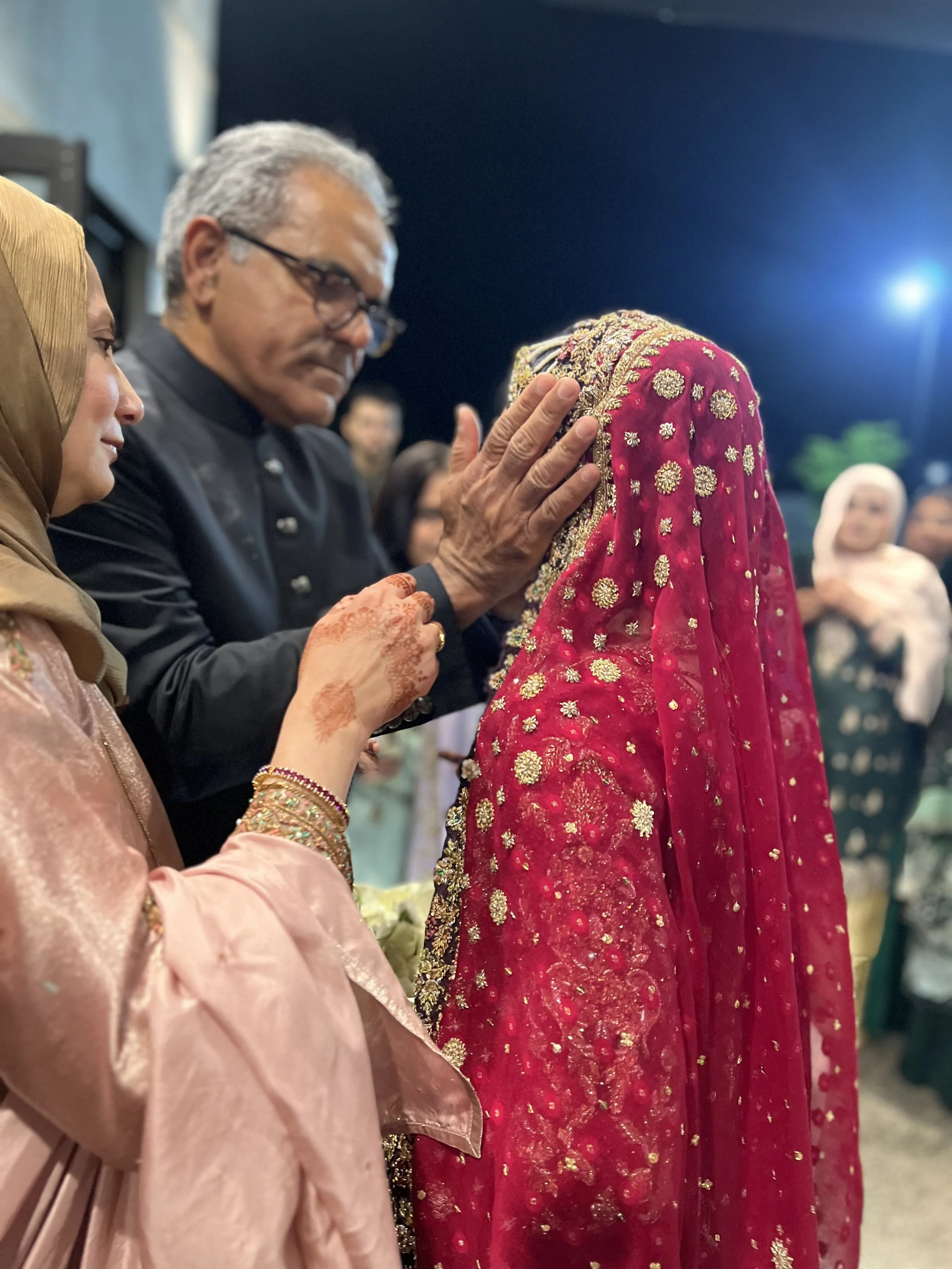 A man with glasses gently touching the forehead of a woman dressed in a red, heavily embroidered traditional Indian wedding outfit. The woman wears a bridal veil and jewelry, and another woman nearby in a pink outfit observes the moment. The scene ap
