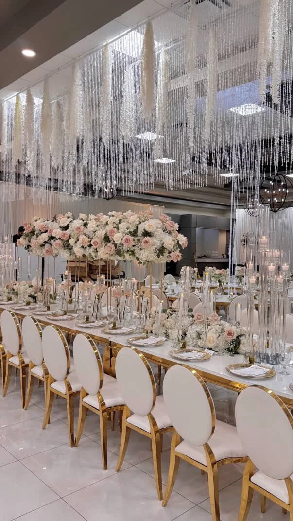Elegant banquet table set for a wedding or celebration with white and pink floral centerpieces, white tableware, gold chairs, and decorative hanging beads and crystals.
