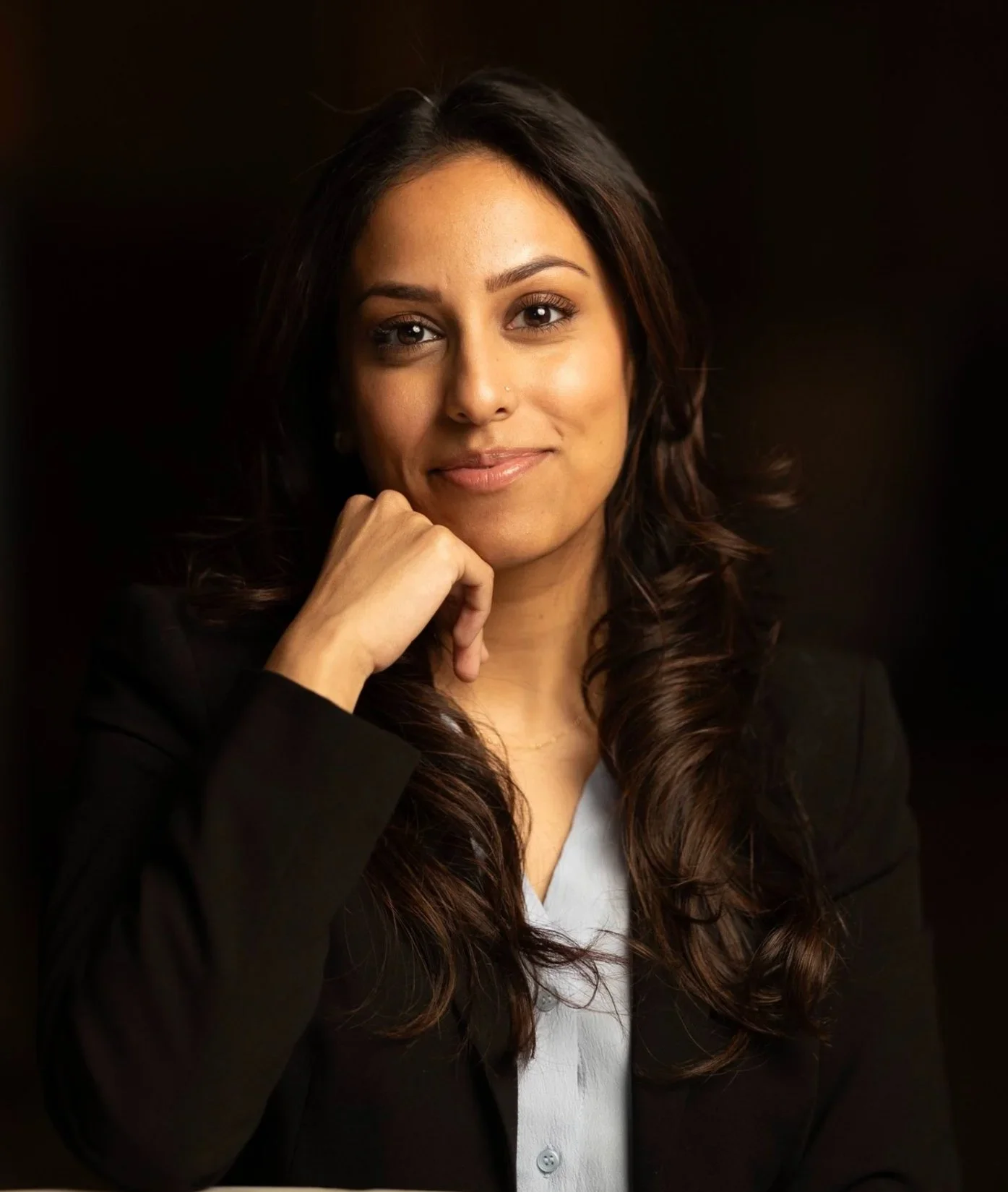 A woman with long wavy hair, wearing a black blazer and light-colored shirt, resting her chin on her hand, smiling softly against a dark background.