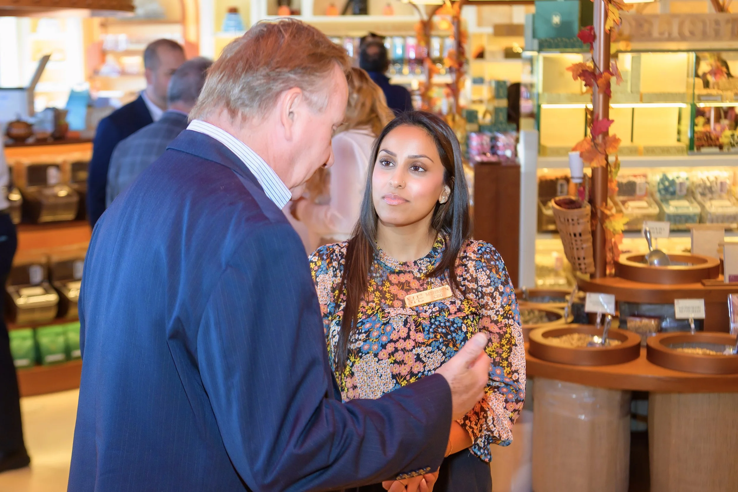 A man and a woman engage in a conversation at a store, with shelves of various products in the background.