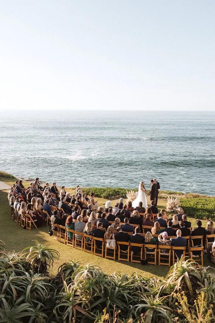Outdoor wedding ceremony with a bride and groom standing before guests on a grassy area overlooking the ocean, with seating and decorative plants.