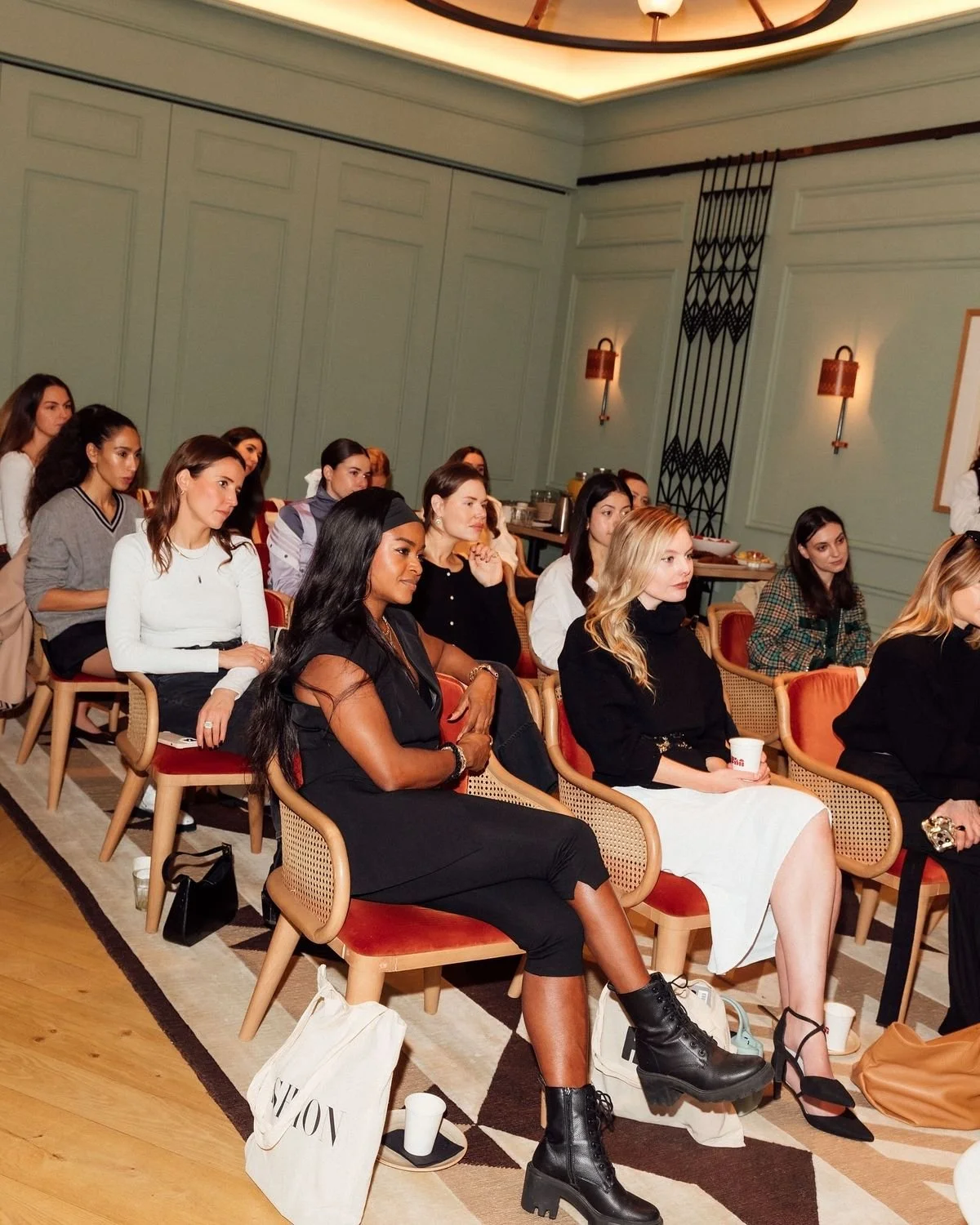 Women attending a conference or seminar in a well-lit room with green walls and wooden floors, seated on red and beige chairs, some holding cups, with a refreshment table in the background.