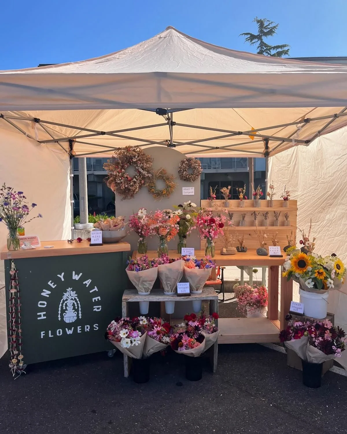 A flower stall called Honey Water Flowers at an outdoor market, displaying colorful bouquets of fresh flowers in vases and buckets, with a decorative wreath on the wall behind and signs with prices. A large sunflower arrangement is on the right, and 