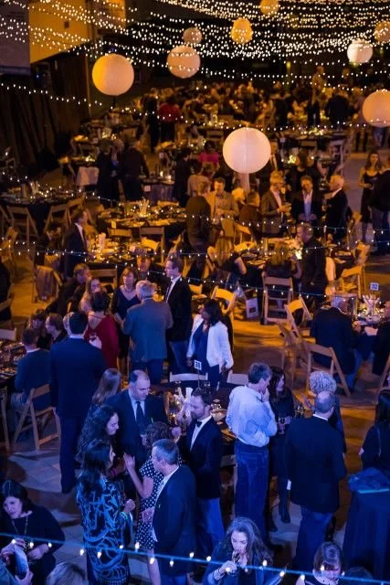 People mingling and socializing at a festive indoor event with hanging string lights and large paper lanterns.