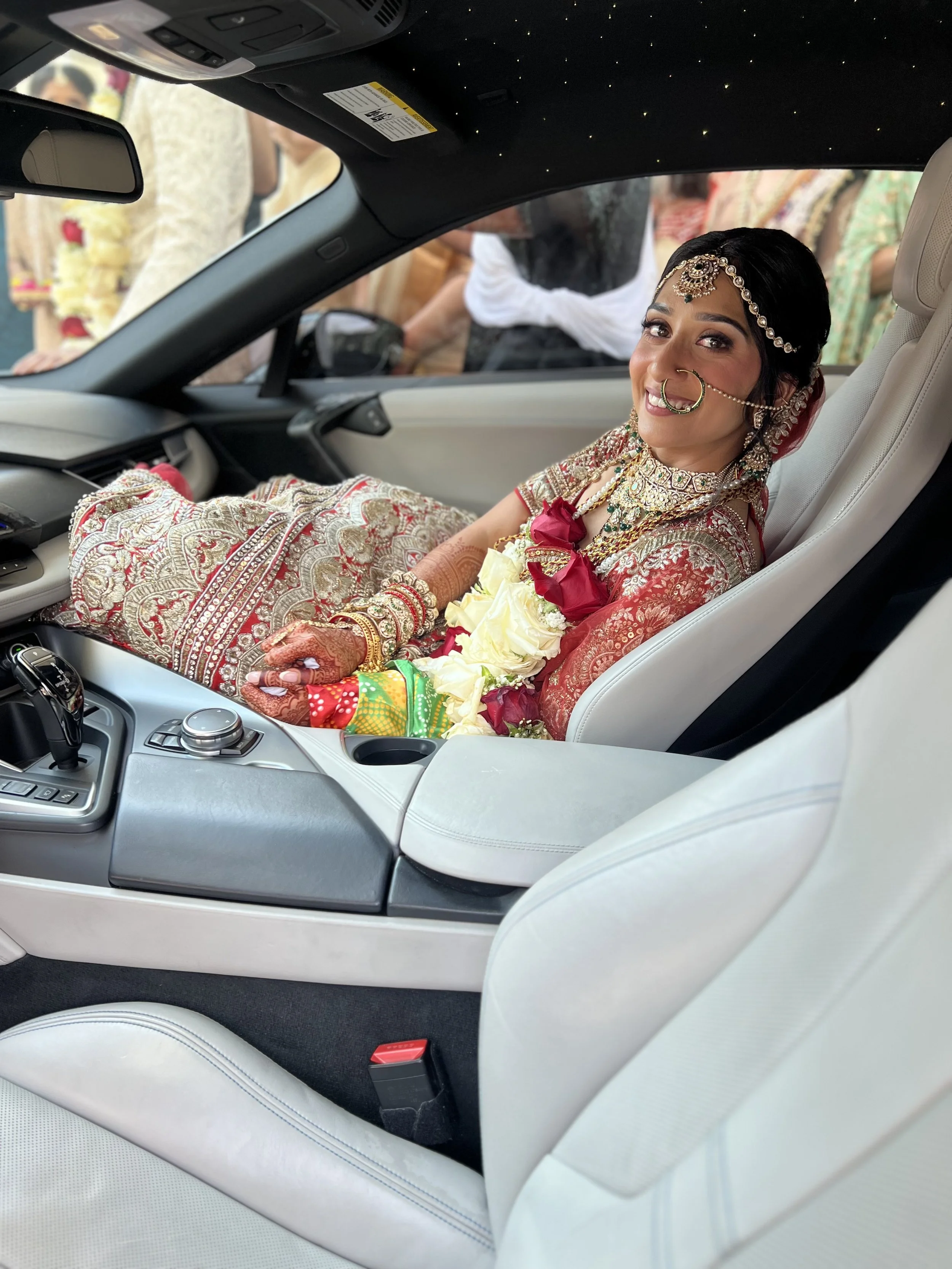 A bride dressed in traditional Indian wedding attire, adorned with jewelry, sitting in a luxury car, smiling with a wedding garland around her neck.