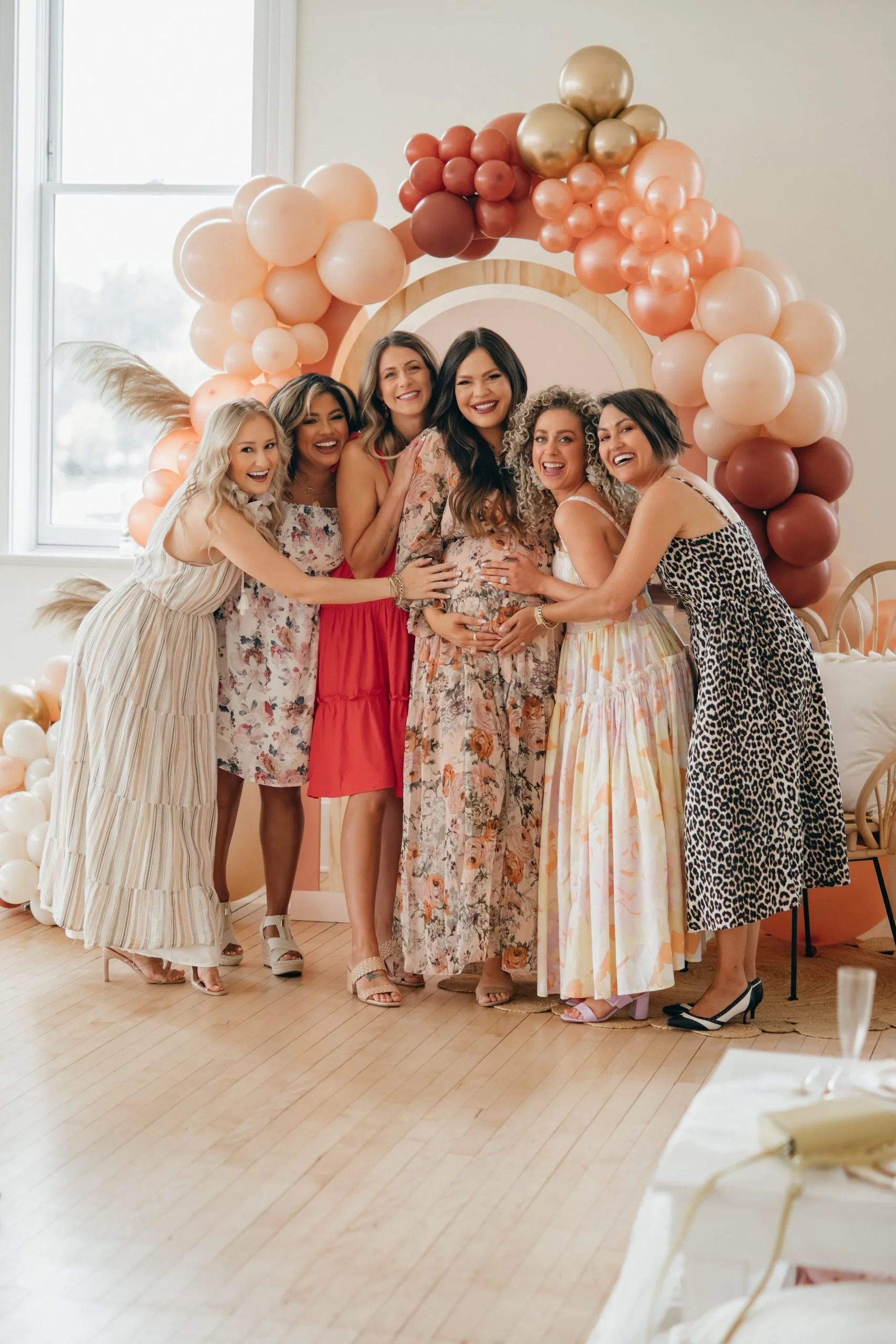 Group of women smiling and hugging a pregnant woman at a celebration, with pink and gold balloons in the background.