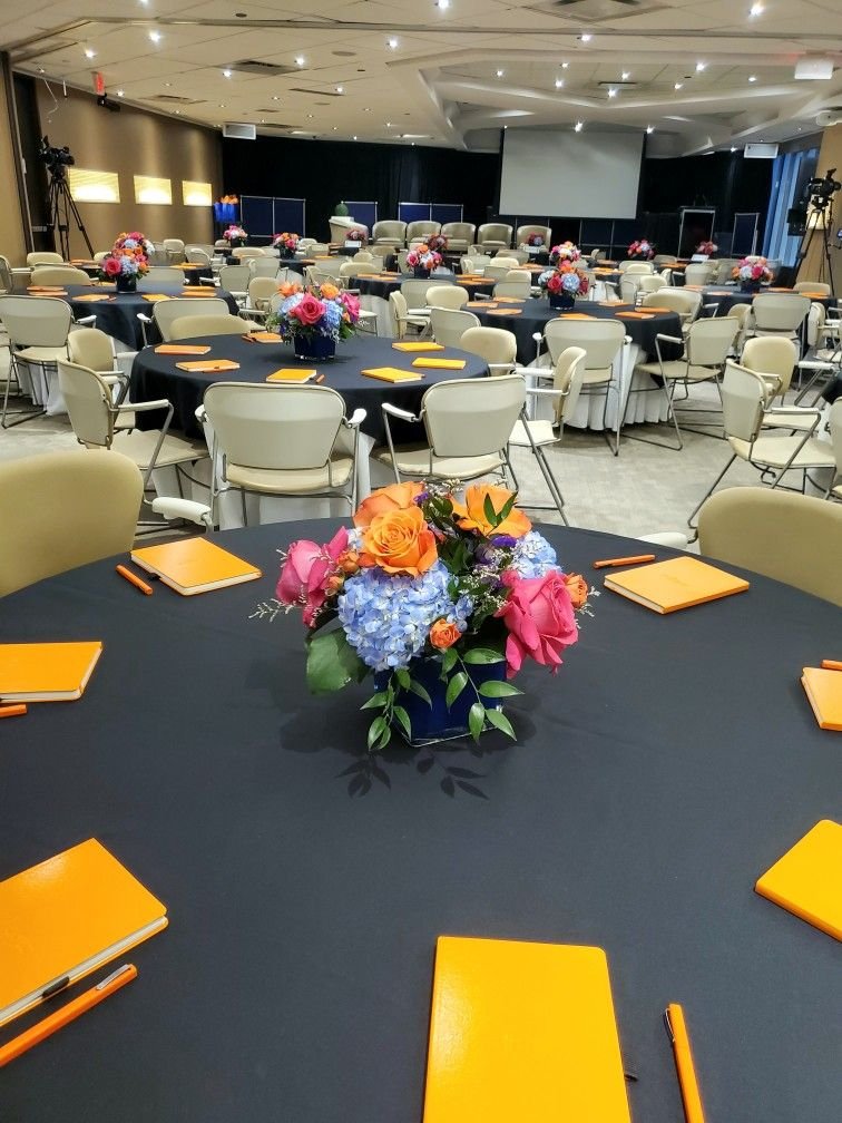 Conference or event room with round tables covered in black tablecloths, each decorated with a colorful flower arrangement. Chairs surround the tables, and there are yellow notebooks with orange pens on each table. A large screen and camera equipment