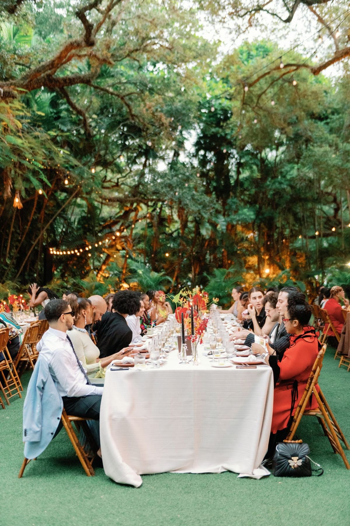 People seated at a long banquet table outdoors, surrounded by lush green trees and string lights, preparing to dine together.