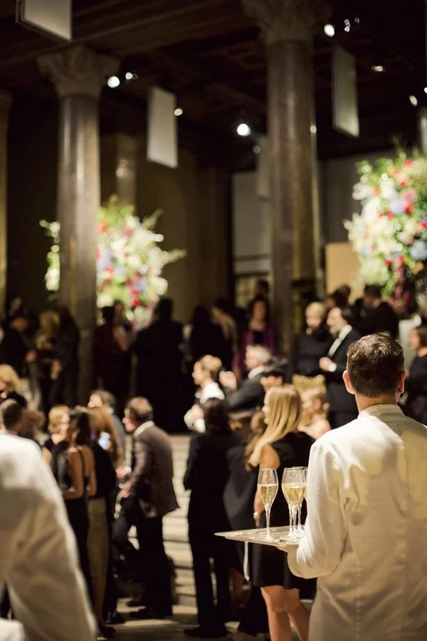 A formal gathering or party with people dressed in elegant attire, standing in a grand venue with tall wooden columns and floral decorations, some holding drinks.