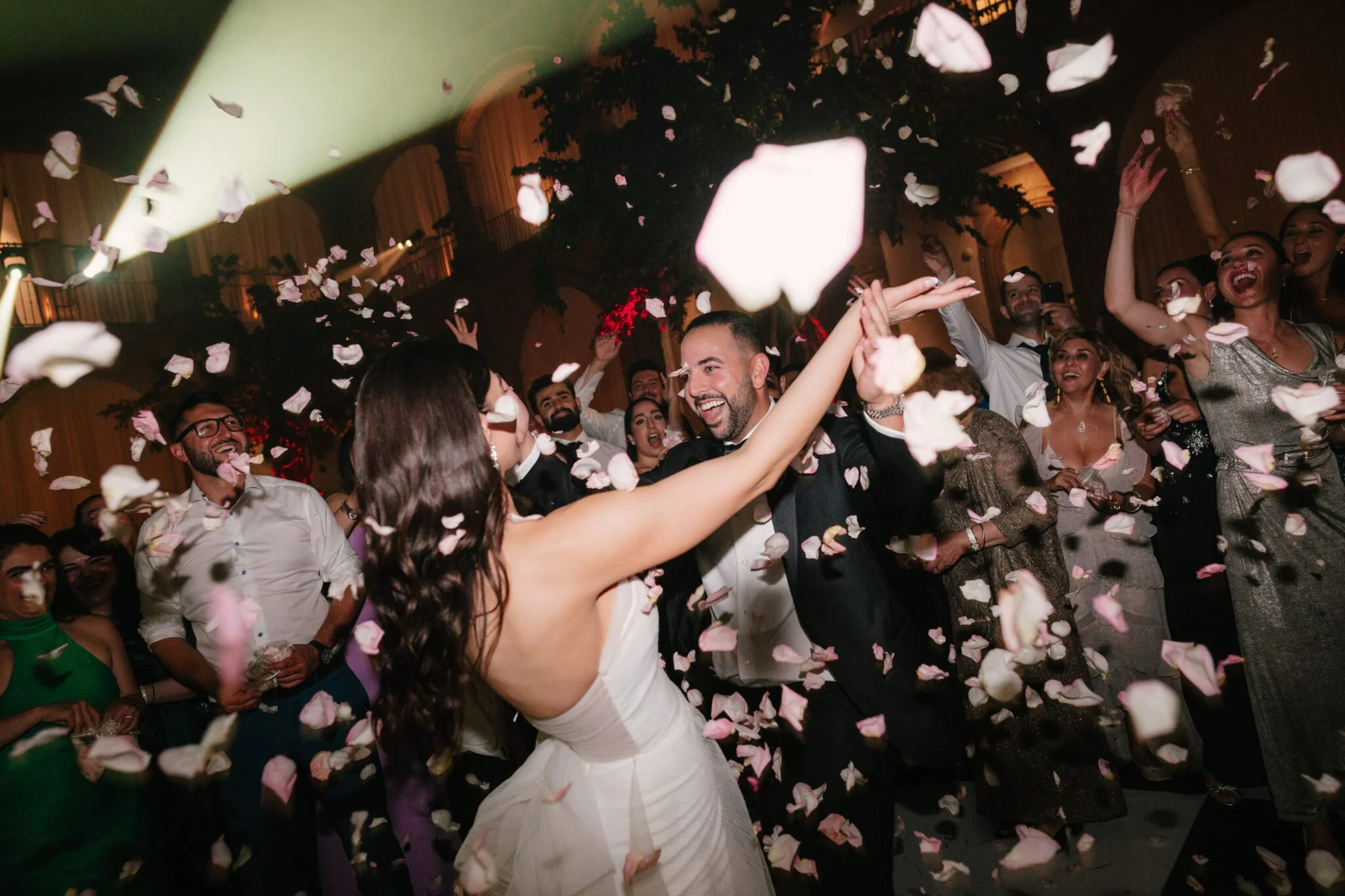 Couple dancing at a wedding reception surrounded by guests with falling rose petals and colorful lighting.