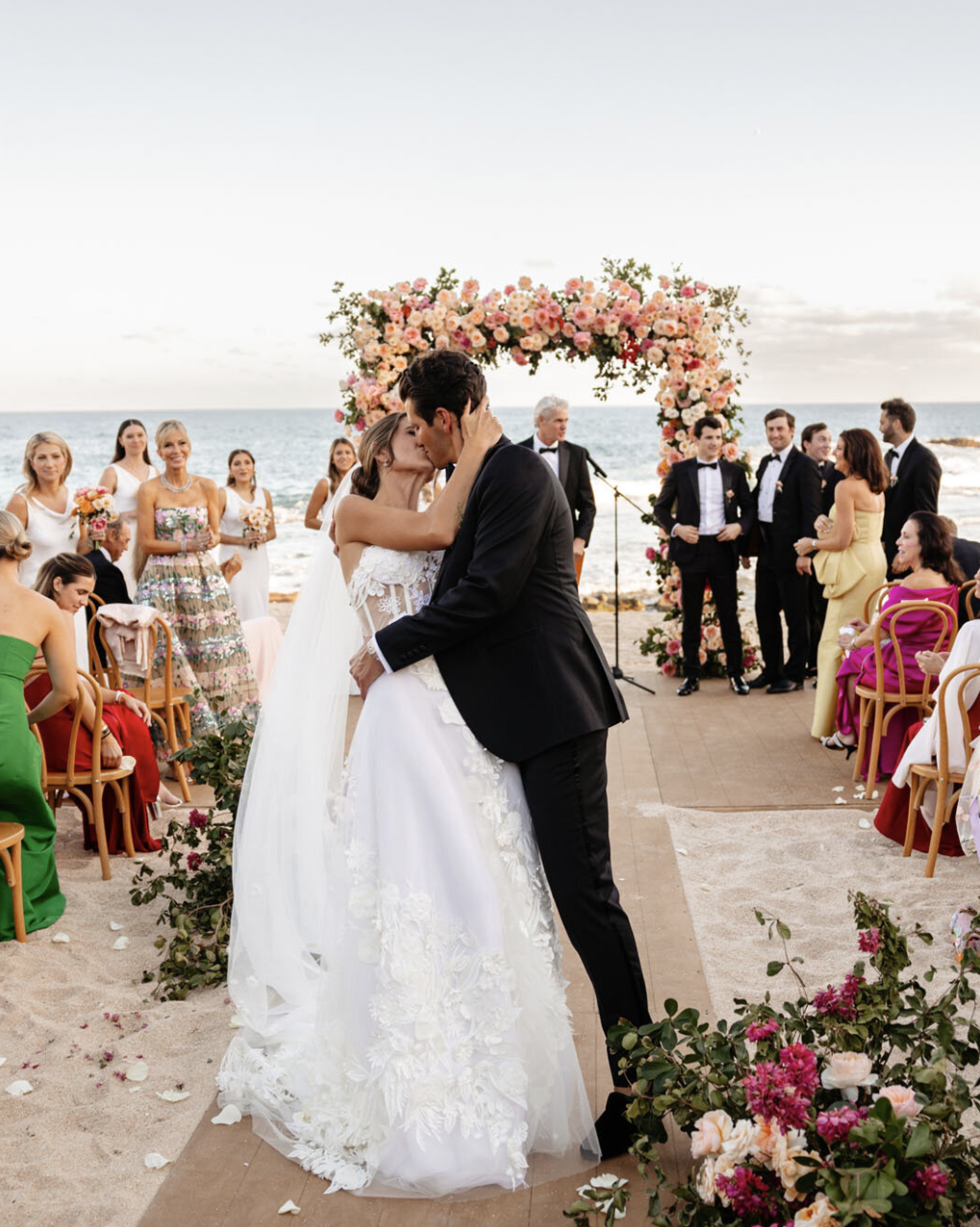 A couple shares a kiss during a beach wedding ceremony, with guests and a floral arch in the background under a partly cloudy sky.