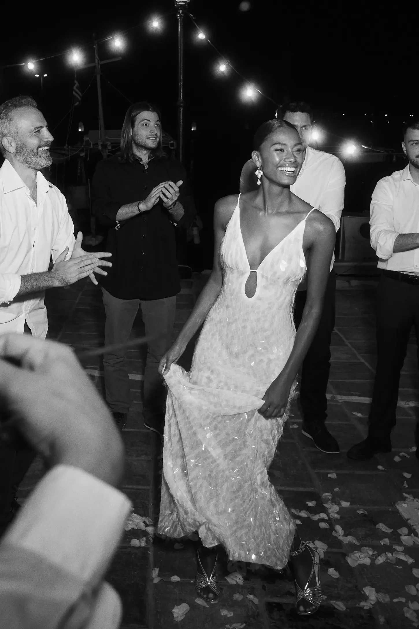 A woman in an elegant, sleeveless dress walking on a dance floor at night, surrounded by people clapping and smiling, with string lights overhead.