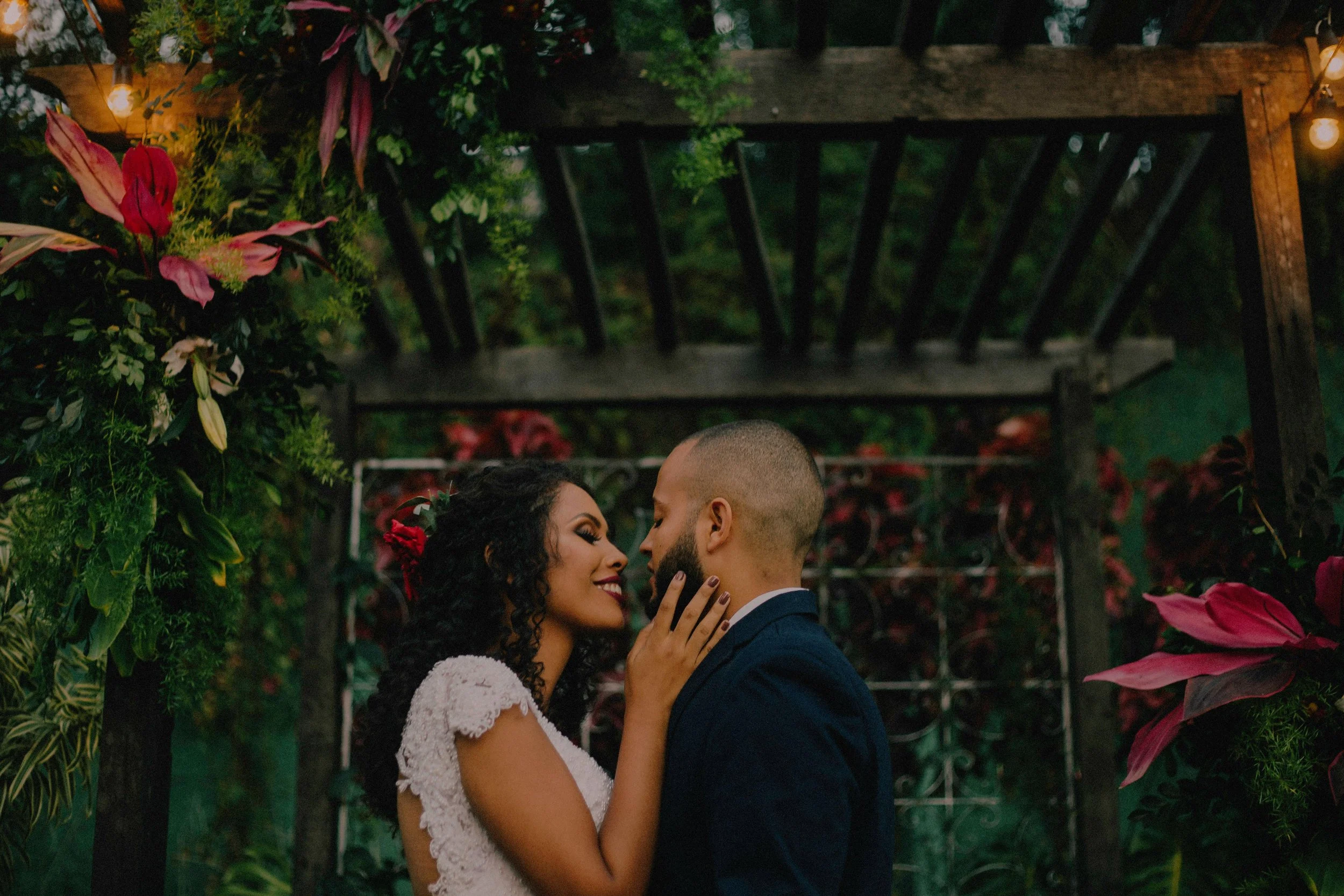 A couple is closely facing each other, touching noses and smiling, amidst lush greenery and floral decorations with hanging lights in the background.