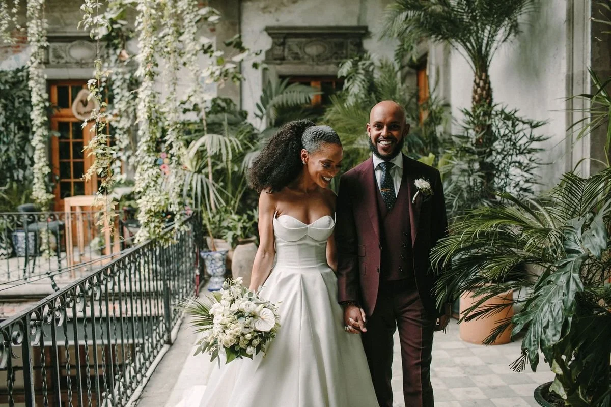 A newlywed couple holding hands and smiling in a lush indoor garden with potted plants, greenery, and natural light.