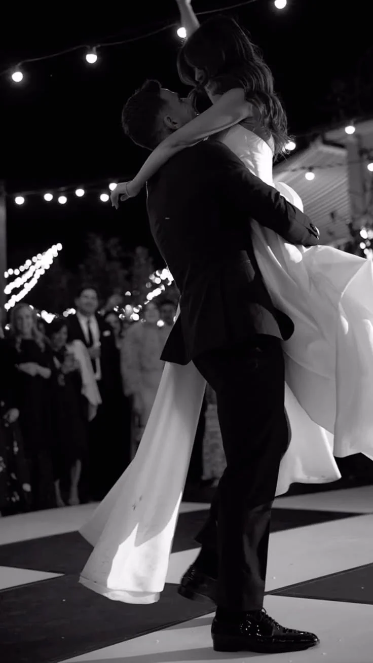 A couple dancing closely at night during an outdoor event with string lights. The man is wearing a suit and the woman is in a long dress, and they appear to be sharing an intimate moment.