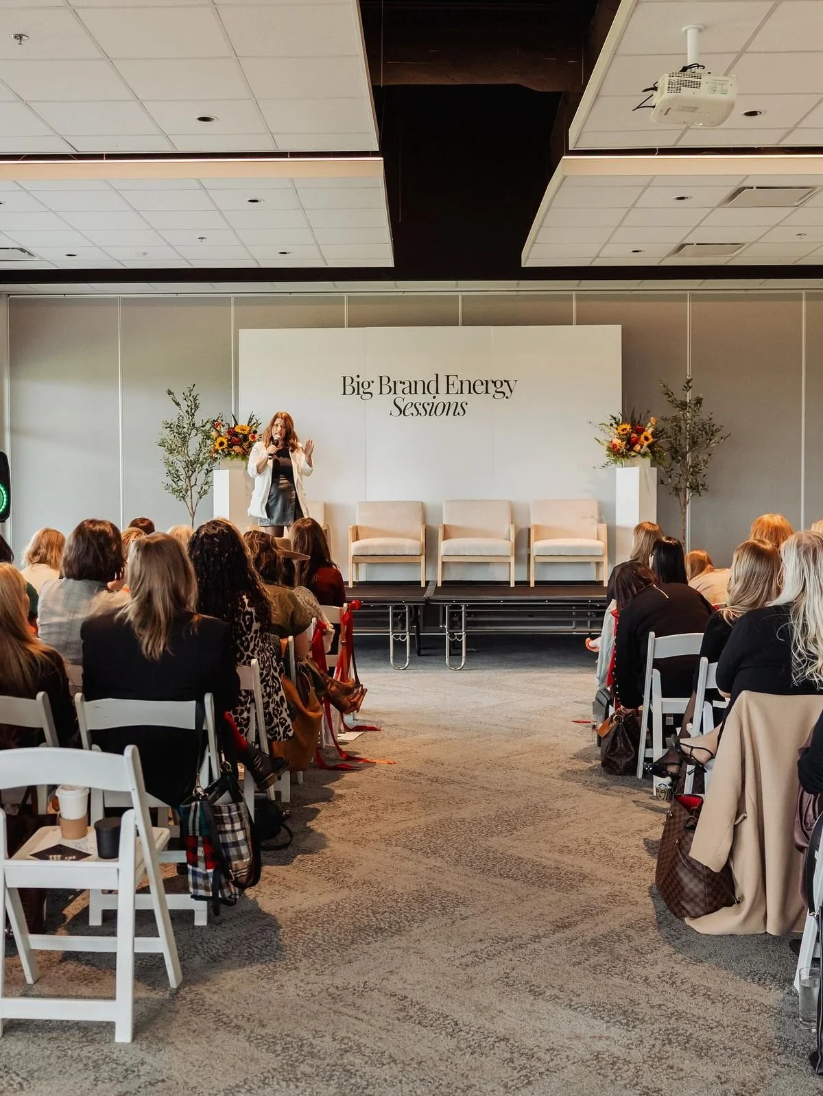A woman speaking on stage at a conference called Big Brand Energy Sessions, with an audience seated in front of her and decorative plants on stage.