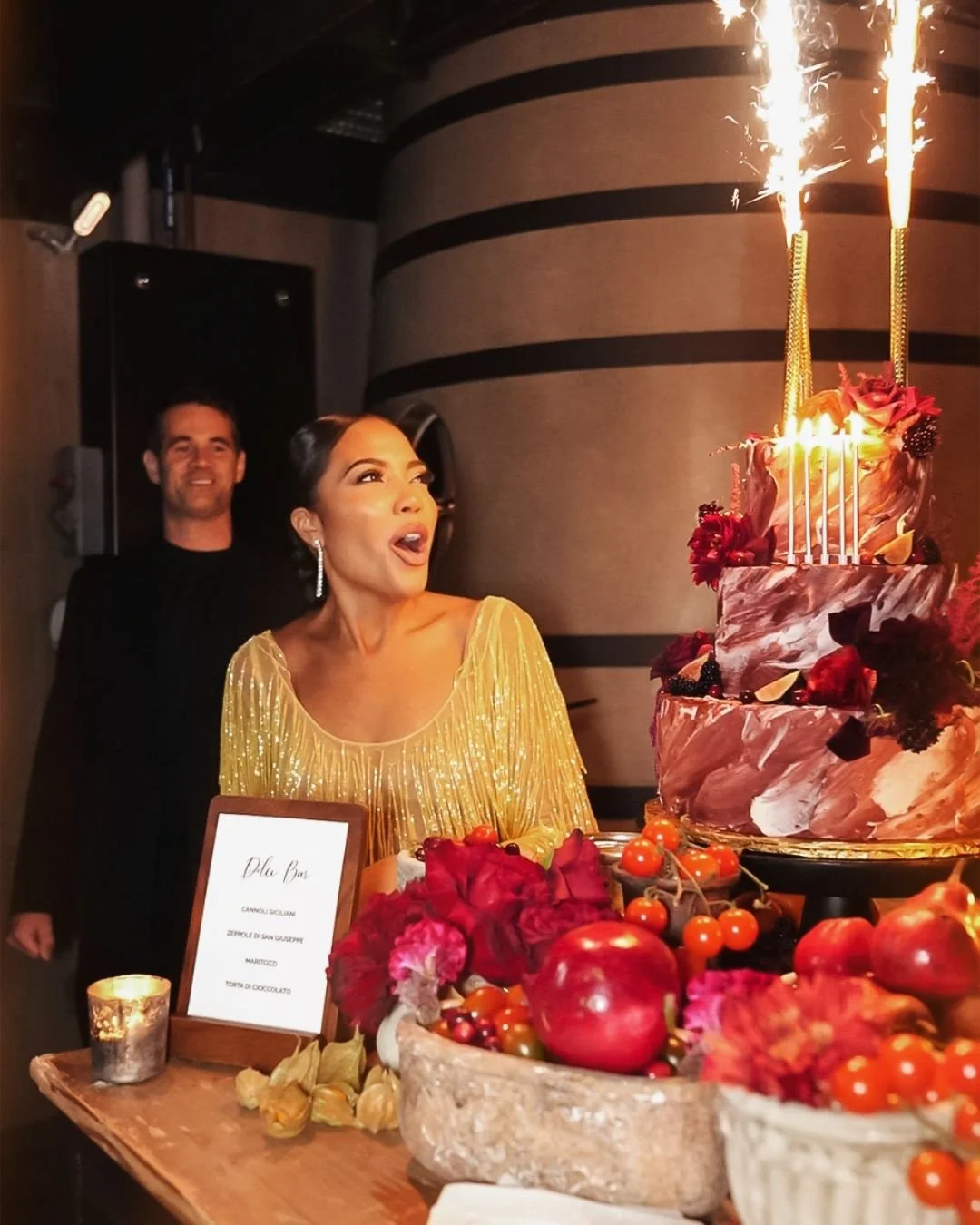 Woman in a gold dress celebrating her birthday with a large decorated cake and sparklers, with a man in black standing behind her in a dimly lit room.