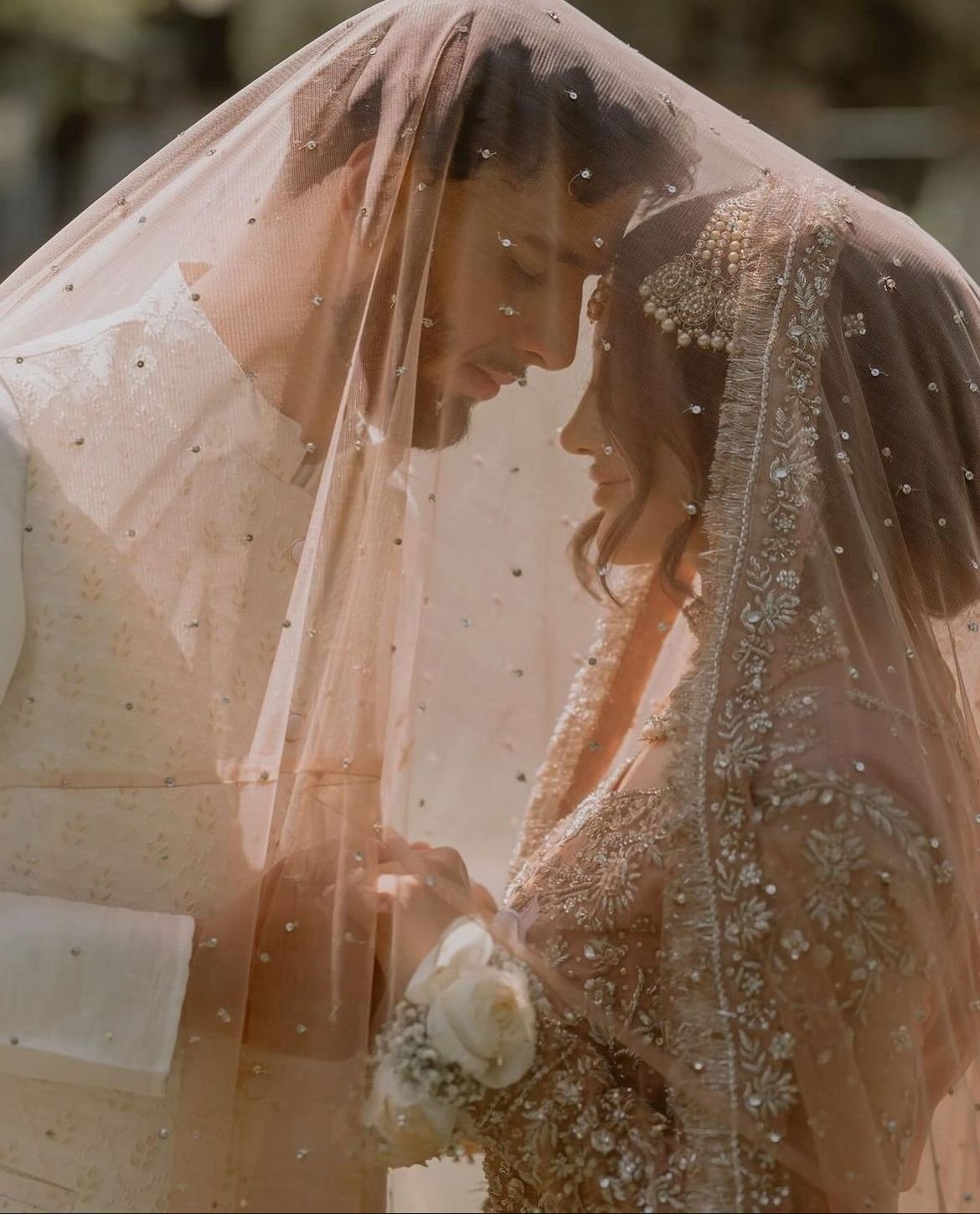 Bride and groom under a sheer veil, gazing at each other, holding hands, with the bride in an embellished dress and intricate headpiece.
