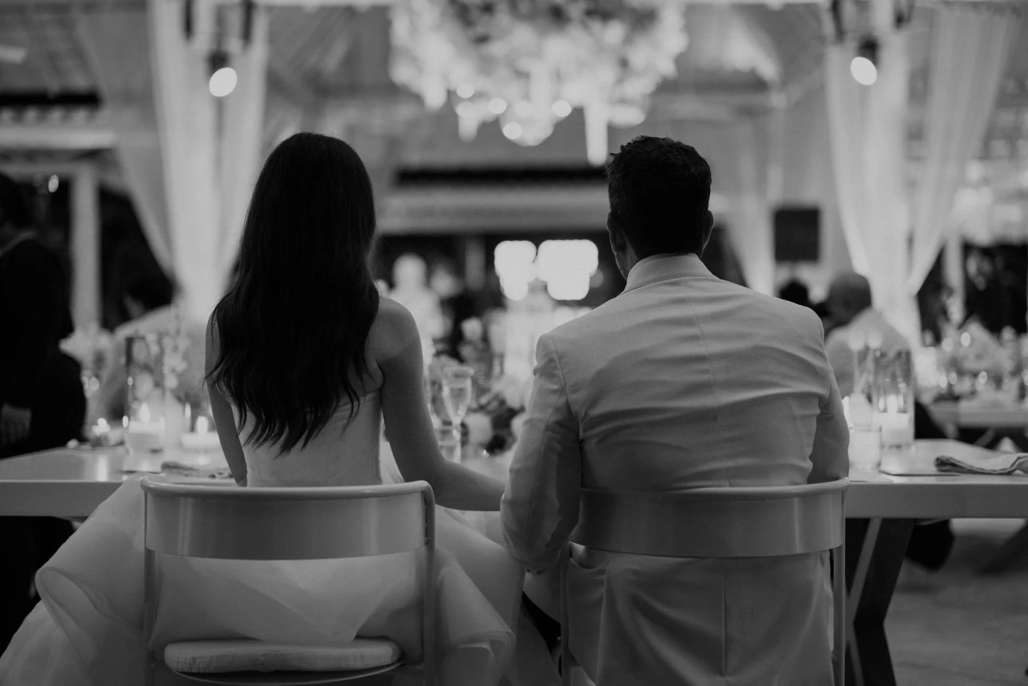 A couple sitting together at a formal event, seen from behind, in a decorated venue with tables and guests.