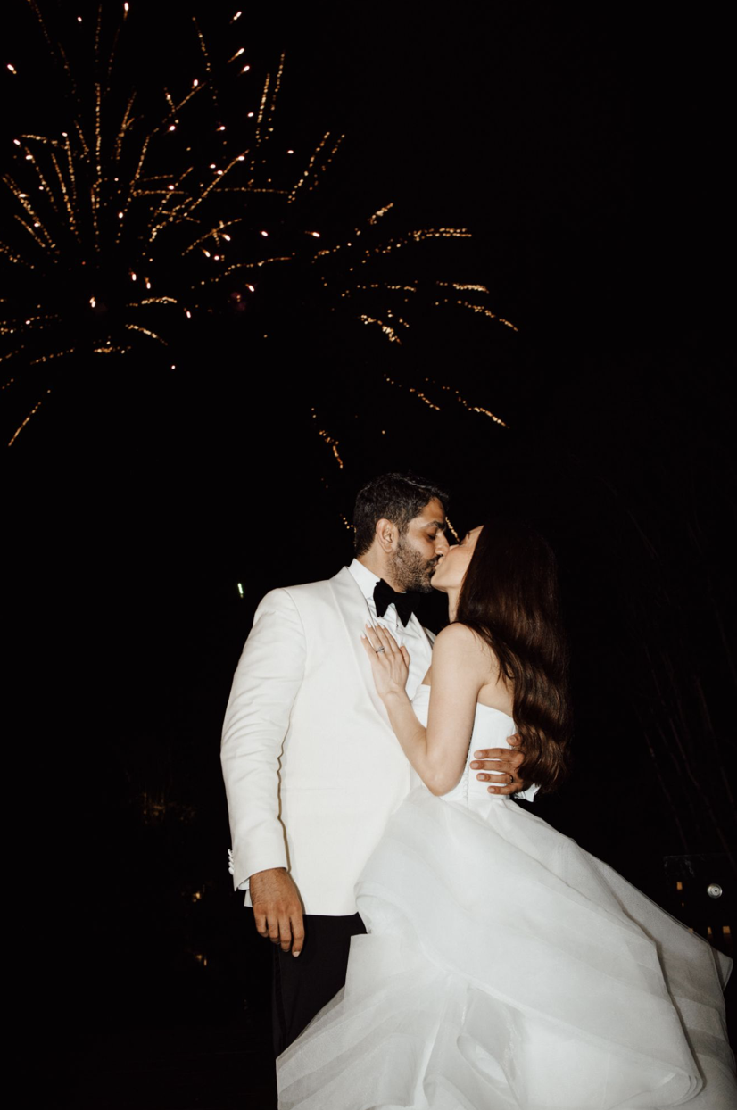 A bride and groom in wedding attire share a kiss during fireworks at night.