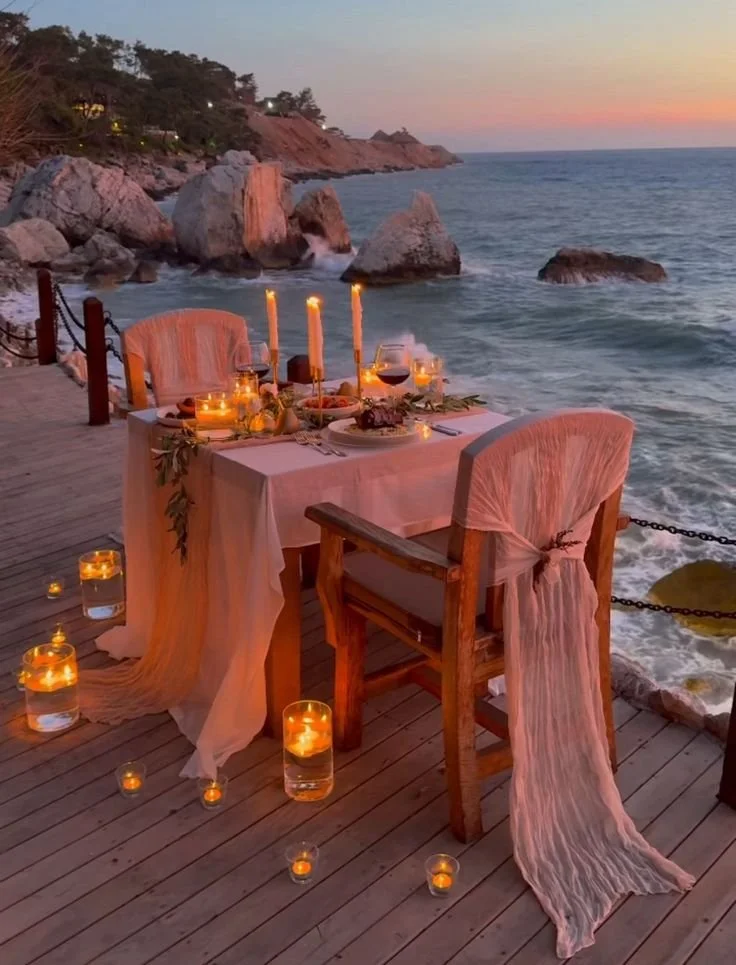 An outdoor dining setup on a wooden deck overlooking the ocean at sunset. The table is decorated with candles and a floral centerpiece, and is set for a meal with plates, glasses, and utensils. The scene is romantic and serene, with rocky cliffs in t