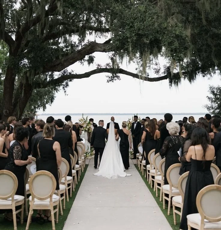 A wedding ceremony taking place outdoors by a lake, with guests seated on both sides of an aisle, under a large tree canopy, with the bride and groom facing each other at the altar.