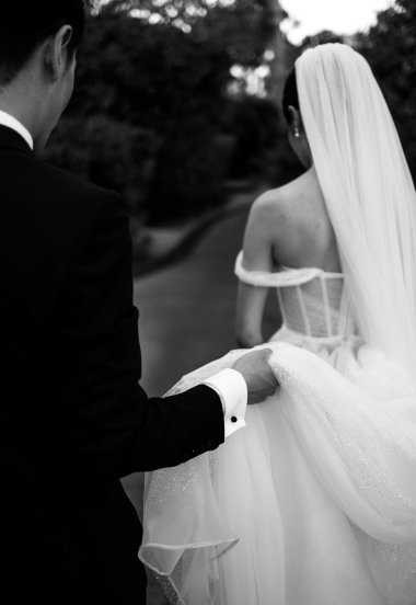 Bride and groom outdoors, bride in wedding dress with veil, groom in tuxedo, holding her dress.