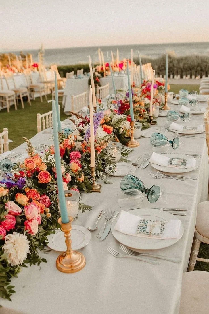 Long outdoor table set for a formal event with flowers, candles, and tableware, near a beach or ocean at sunset.