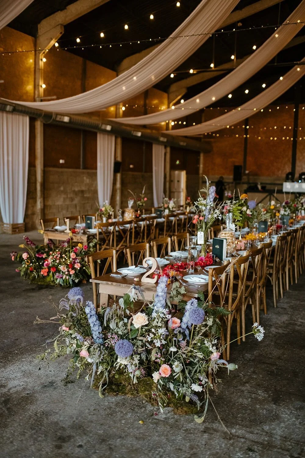 Wedding reception setup with long wooden tables decorated with floral arrangements and tableware, in a rustic venue with draped fabric and string lights overhead.