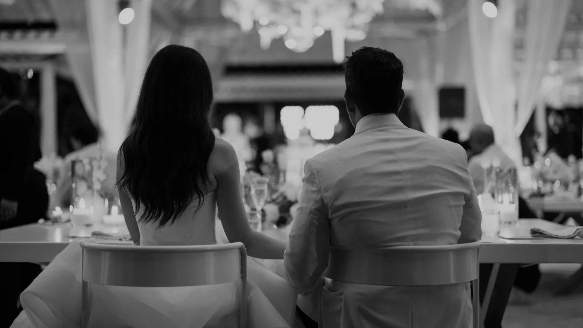 A black-and-white photo of a bride and groom sitting together at a wedding reception, holding hands and facing away from the camera, with other guests and tables visible in the background.