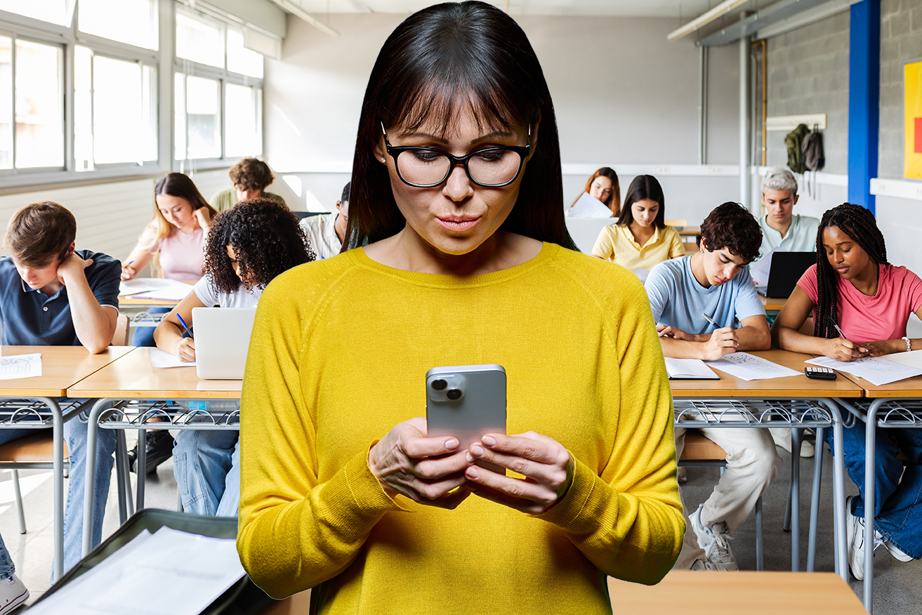 A teacher with glasses wearing a yellow sweater standing in a classroom, looking at her AlertNet advisory on her phone, with students seated at desks in the background.