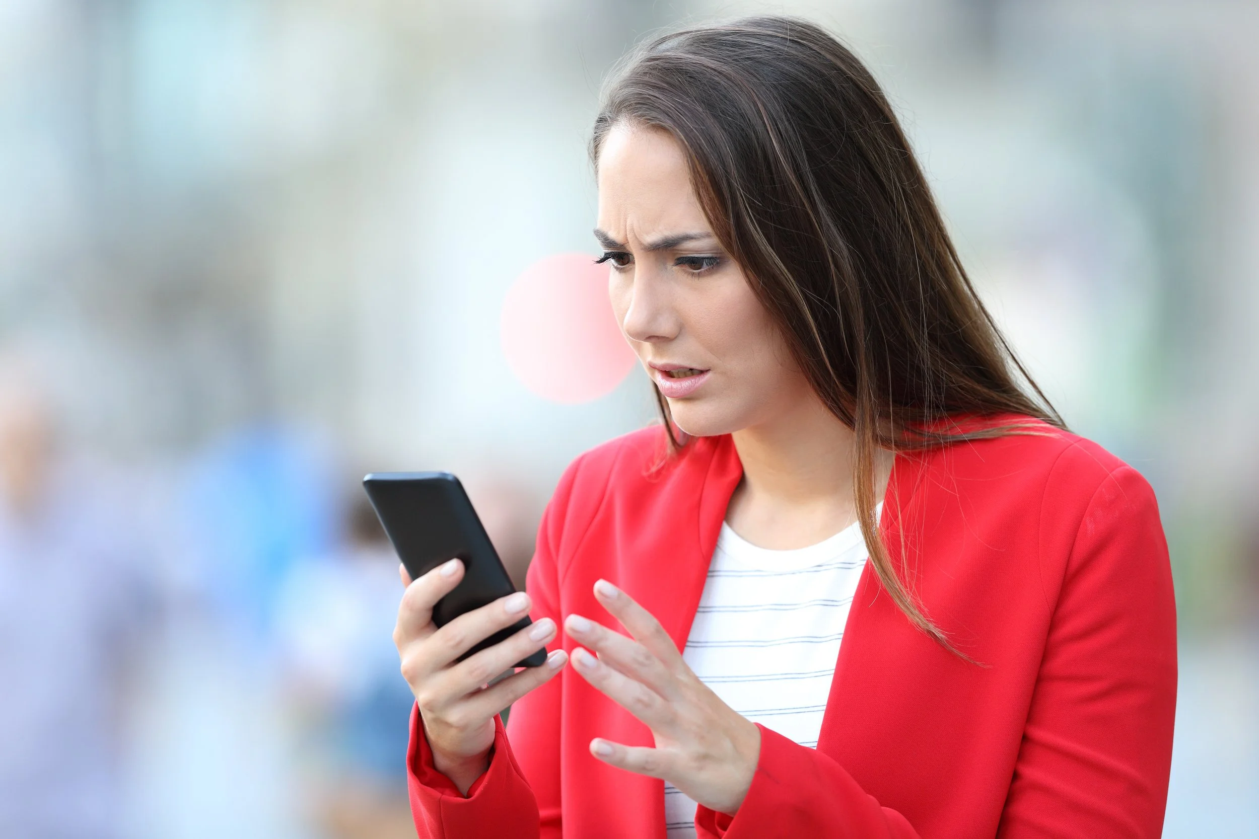 A young woman with long brown hair, wearing a red blazer over a white and gray striped shirt, looking at her smartphone with a confused or concerned expression after she receives an NFSI AlertNet firearm breach advisory