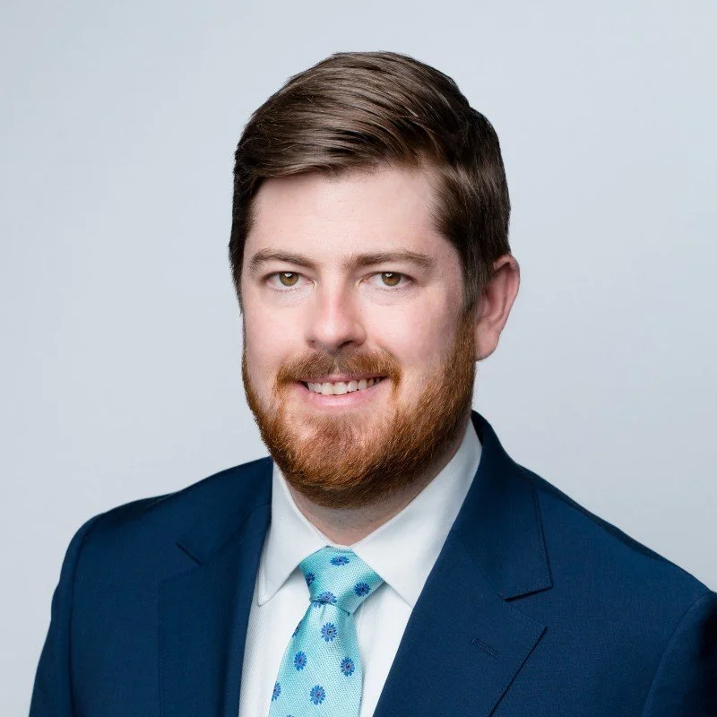 Professional headshot of a young man with brown hair and beard, smiling, dressed in a navy suit, white shirt, and light blue tie with dark blue floral pattern, against a light gray background.
