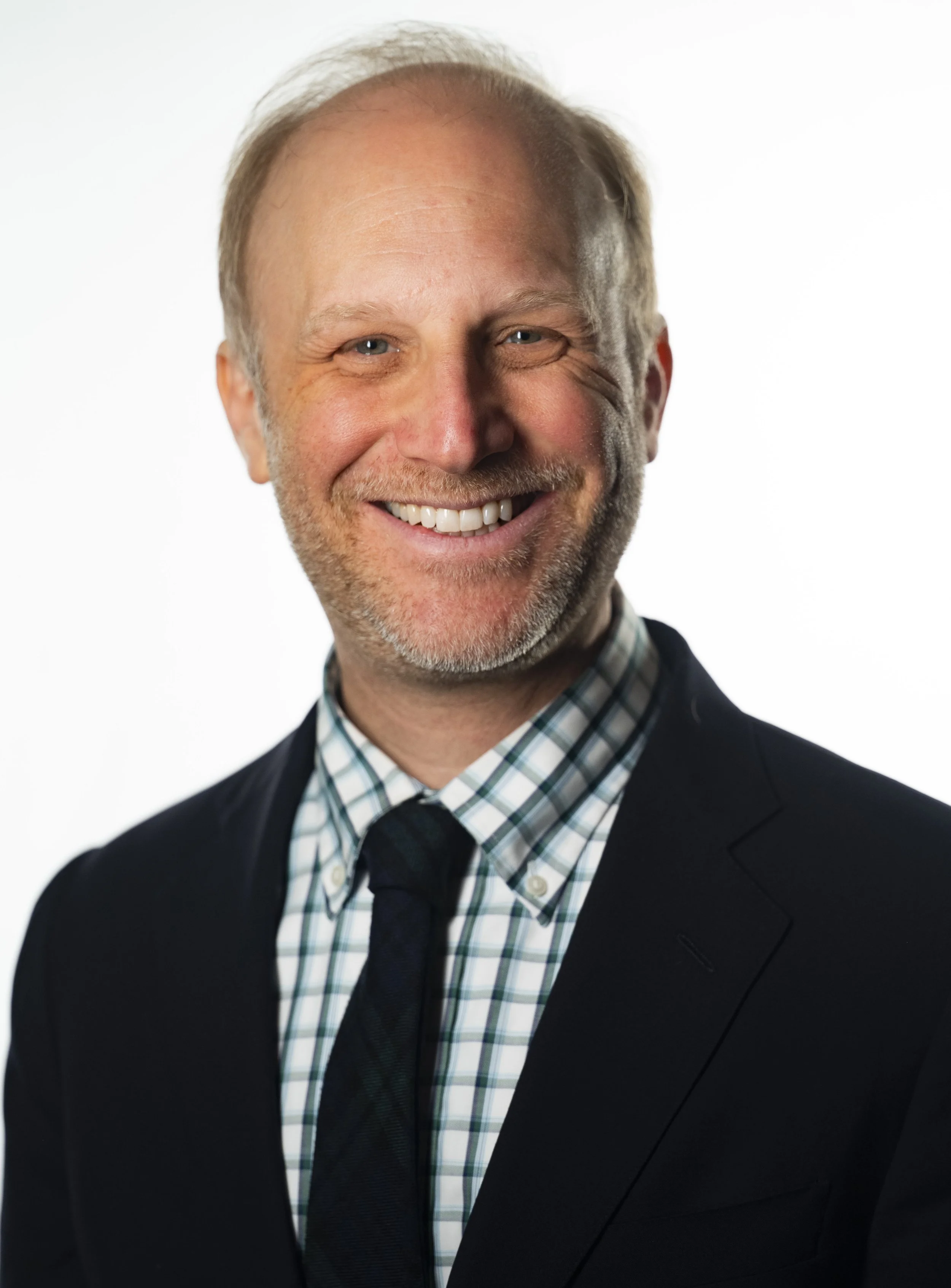 Professional portrait of a smiling middle-aged man with light hair, beard, wearing a dark suit, checkered shirt, and tie, against a plain white background.
