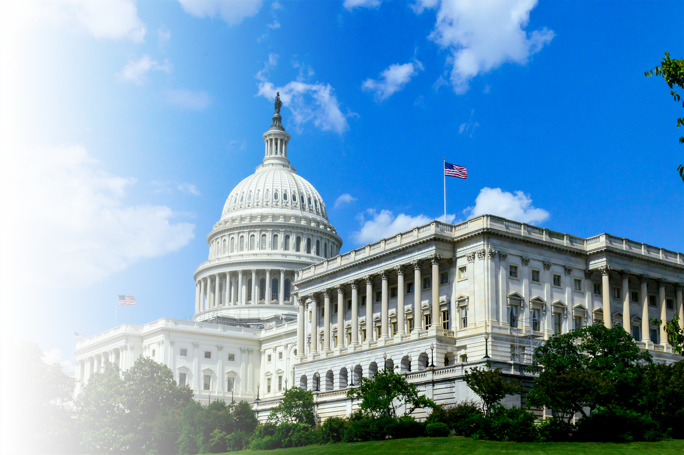 The United States Capitol building in Washington, D.C., with a blue sky and scattered clouds in the background. The building is white with a large dome and American flags on the roof, surrounded by green trees.