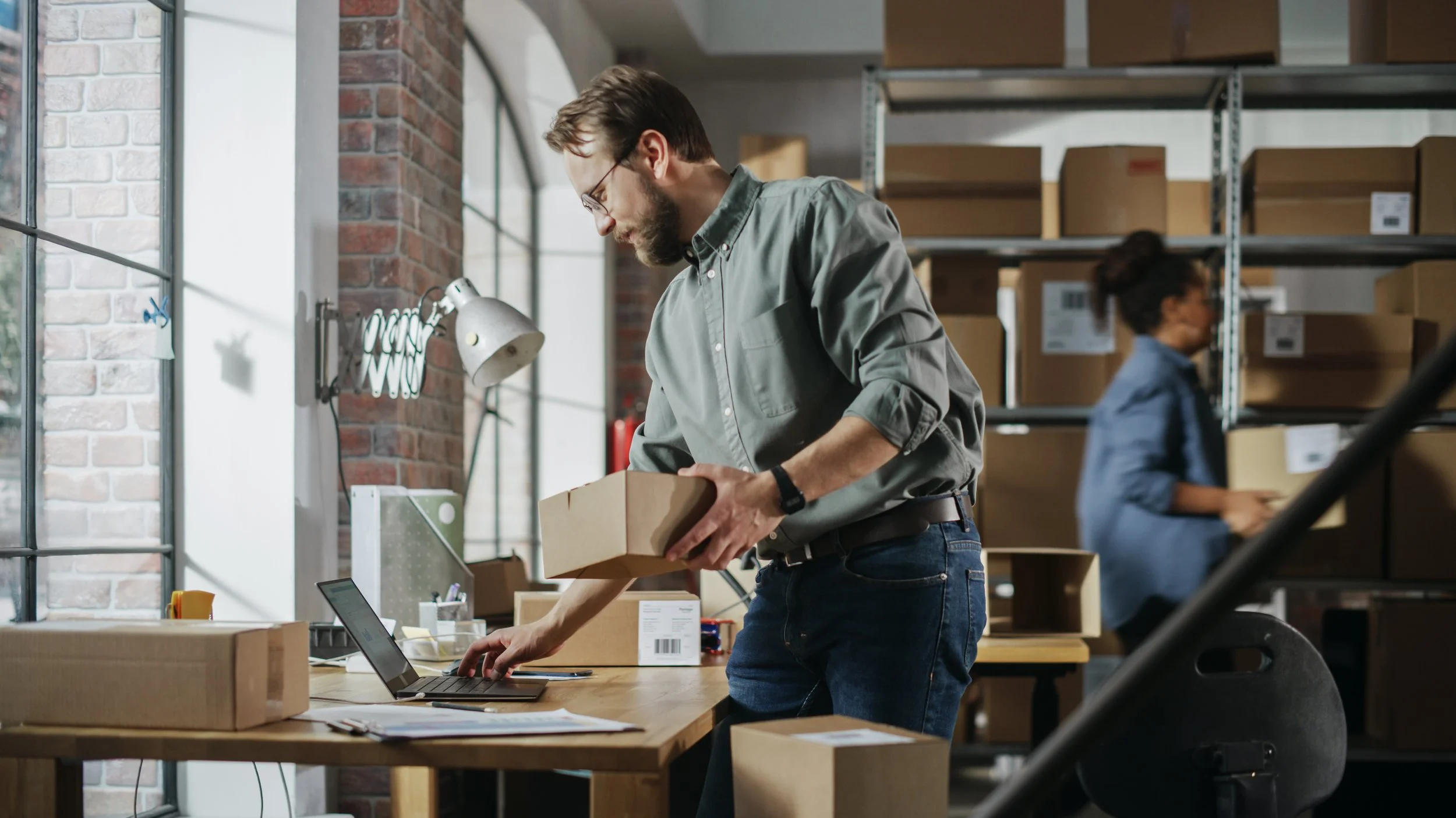 Man in gray shirt and glasses working with a cardboard box at a desk, with a woman in the background handling boxes in a warehouse.
