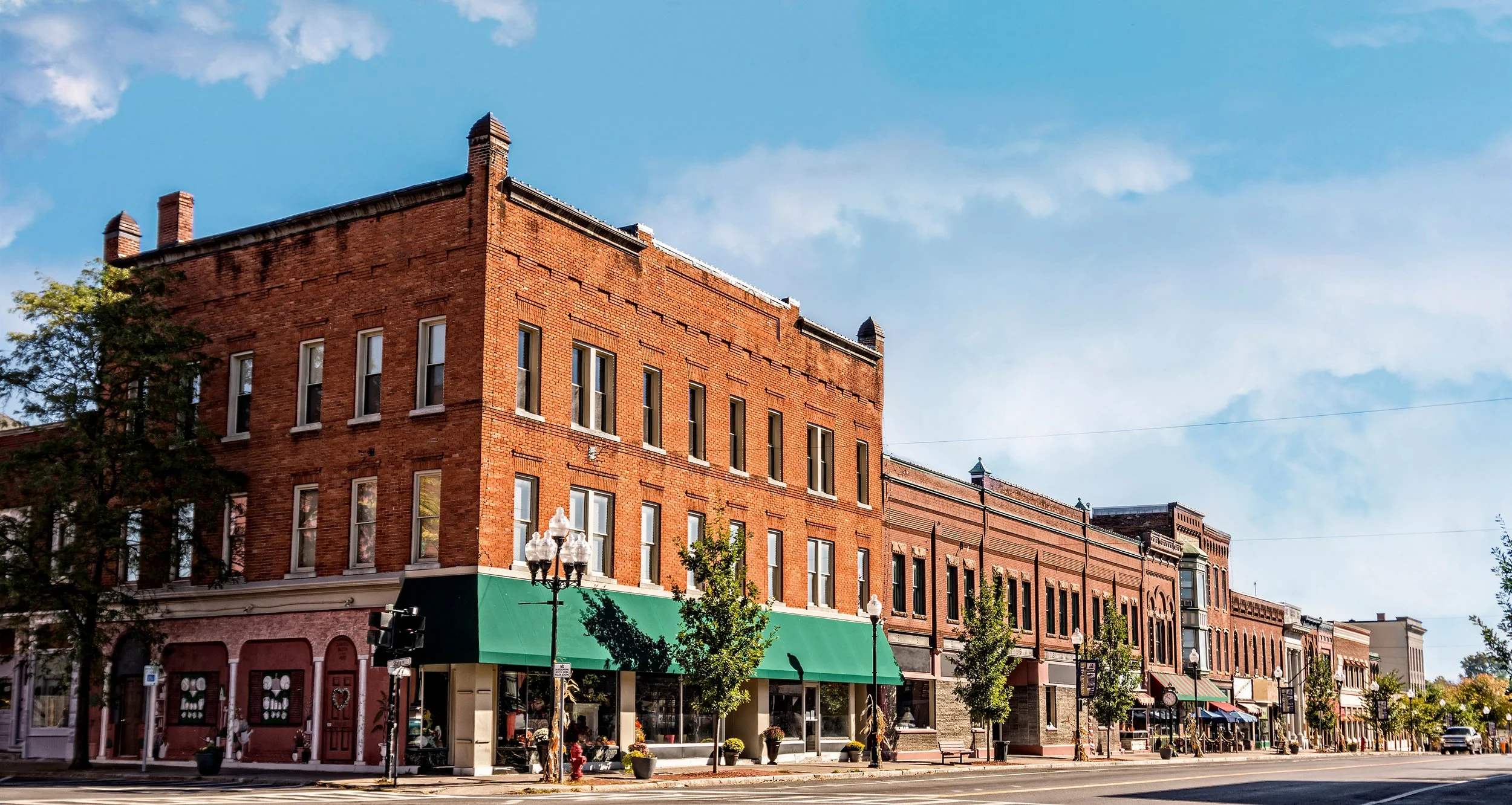 Street view of a small downtown area with brick buildings, storefronts, and trees, under a blue sky.