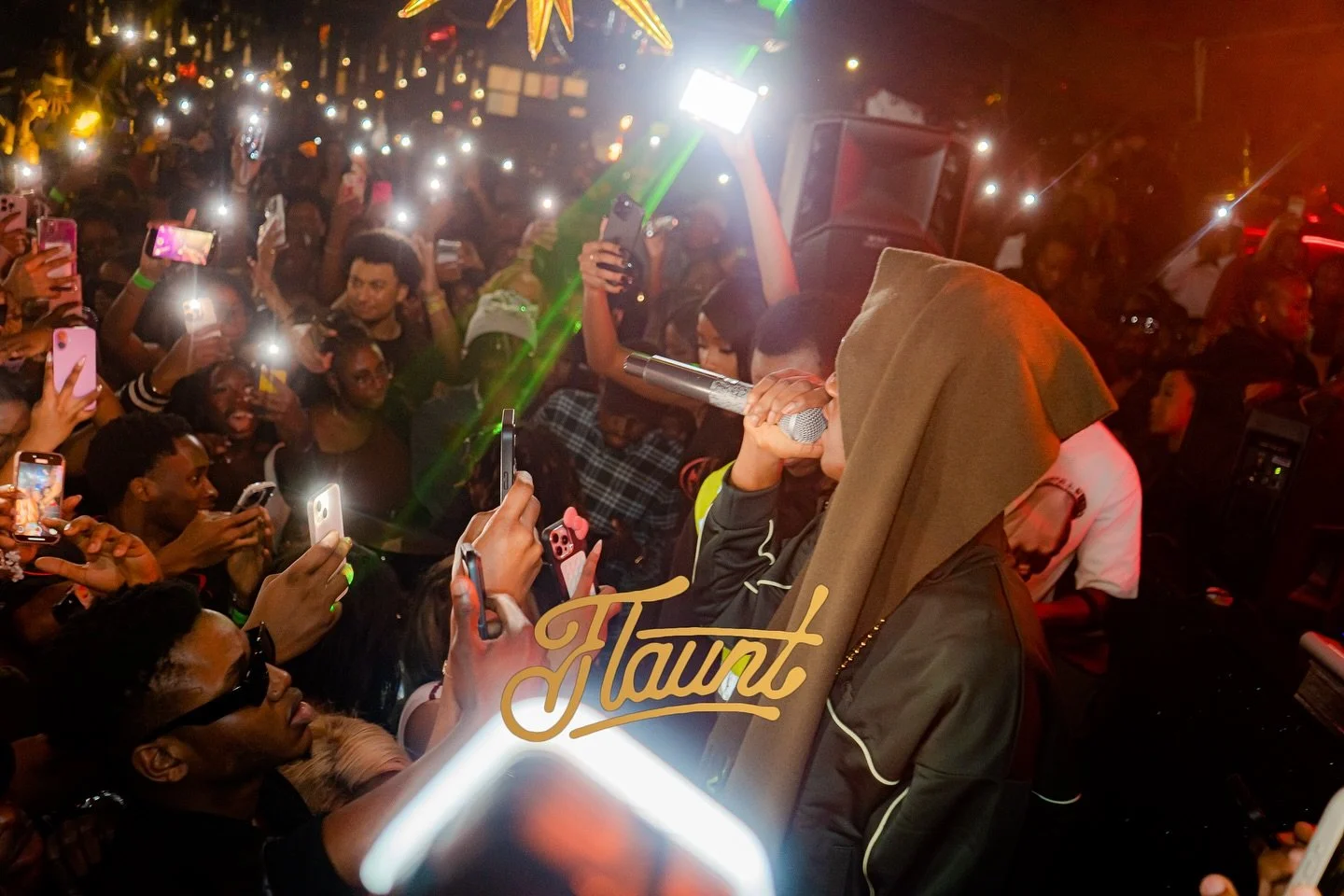 Mavo on the microphone, performing at a crowded flaunt night with many people taking photos and videos.