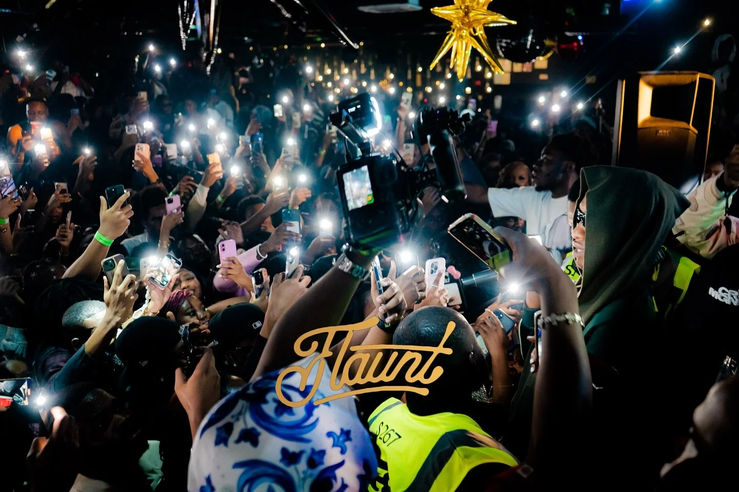 Crowd of people at a flaunt concert, many holding up smartphones and taking photos or videos, with bright lights and a star-shaped decoration hanging from the ceiling.