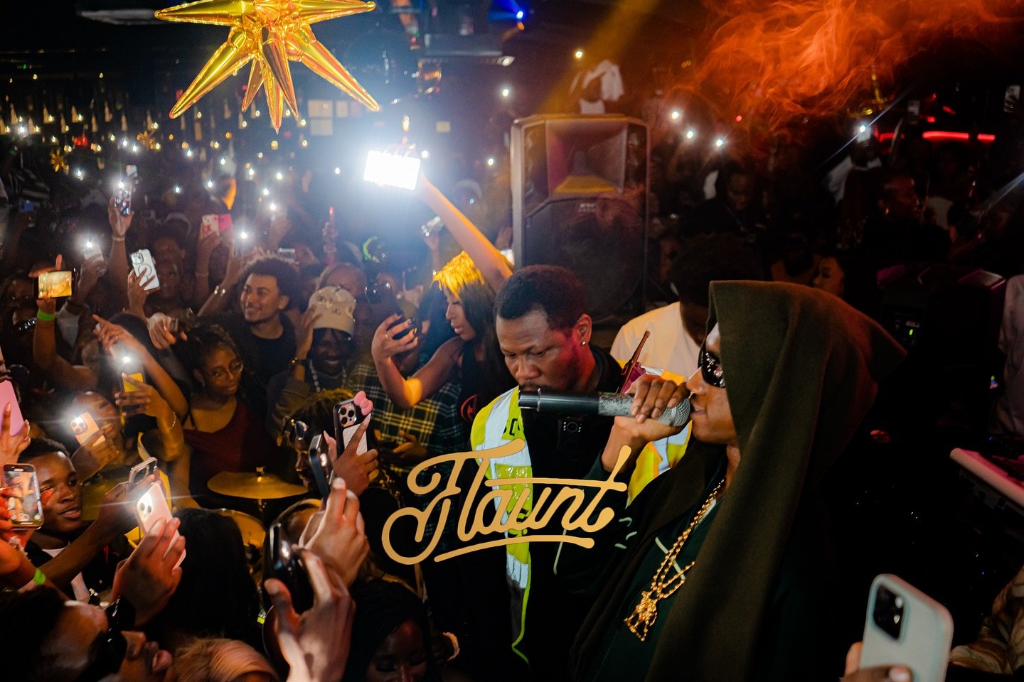 Mavo on the microphone, performing at a crowded flaunt night with many people taking photos and videos.