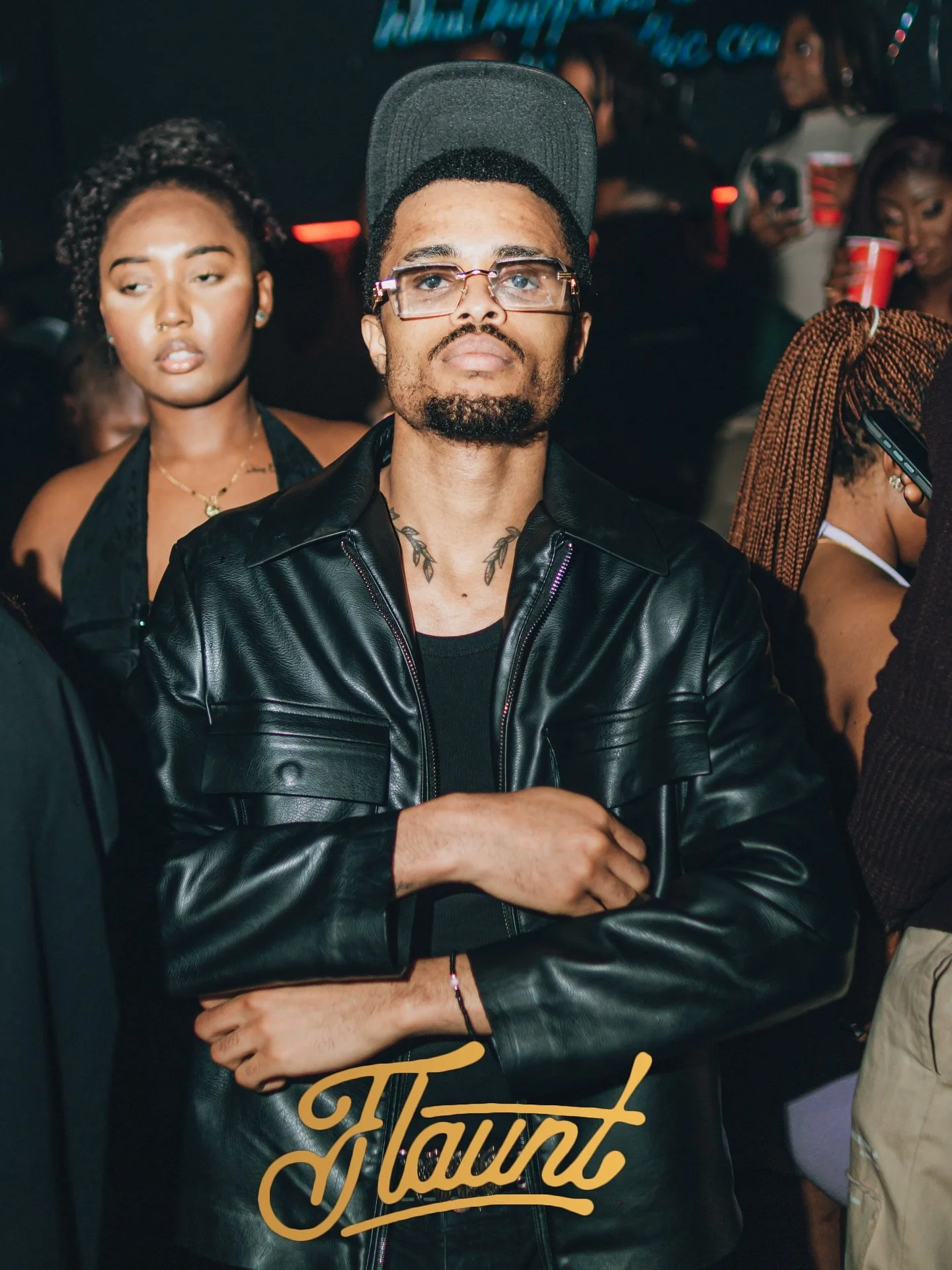 A dapper gentleman with glasses, tattoos, and a cap, wearing a black leather jacket, standing with arms crossed at a flaunt event with his admirers in the background. The word 'Haunt' must have been at the back of their minds.