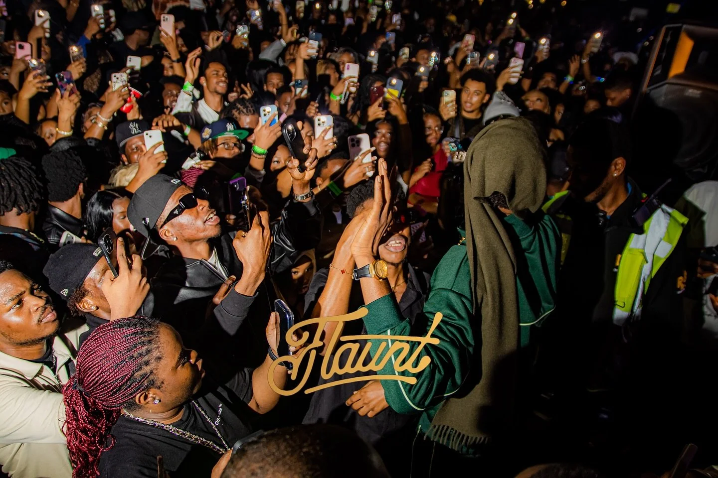 Mavo on the microphone, performing at a crowded flaunt night with many people taking photos and videos with security personnels present.