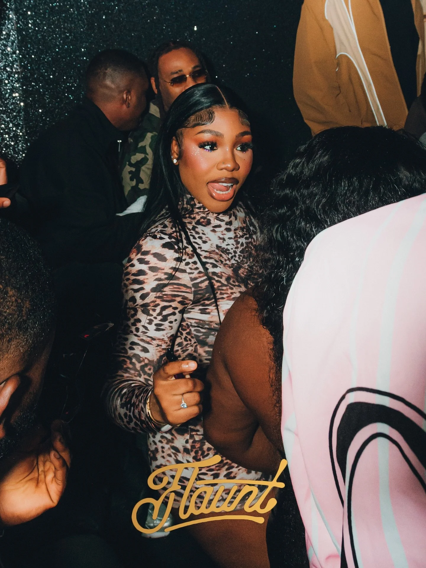 A gorgeous young lady with black hair and makeup, wearing a leopard print top at a flaunt event. She appears to be talking or reacting to something, with many people around her. The background is dark and glittery.
