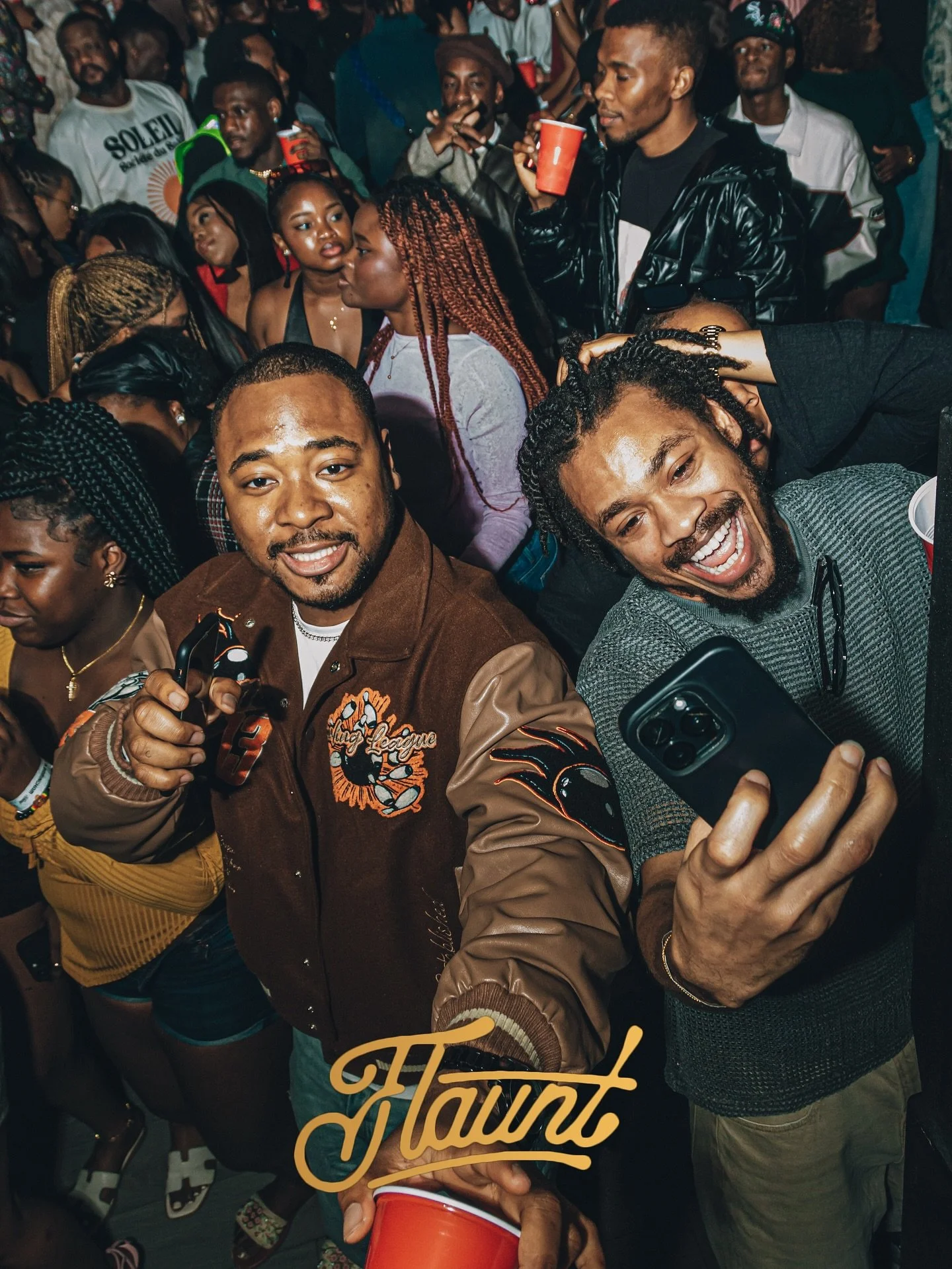 A group of people enjoying a flaunt night, with two men in the foreground taking a selfie and smiling