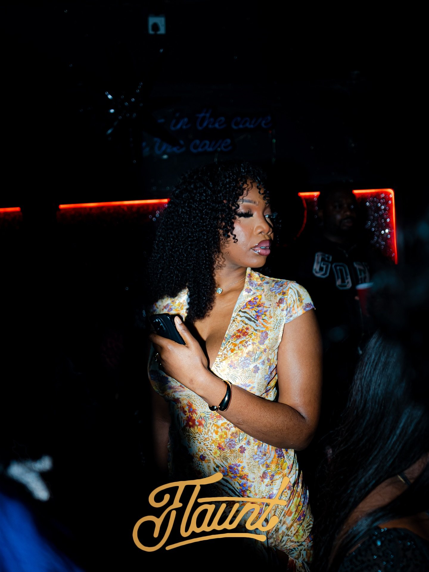 A woman with curly hair wearing a floral dress in a dark  and a man in the background with a drink.