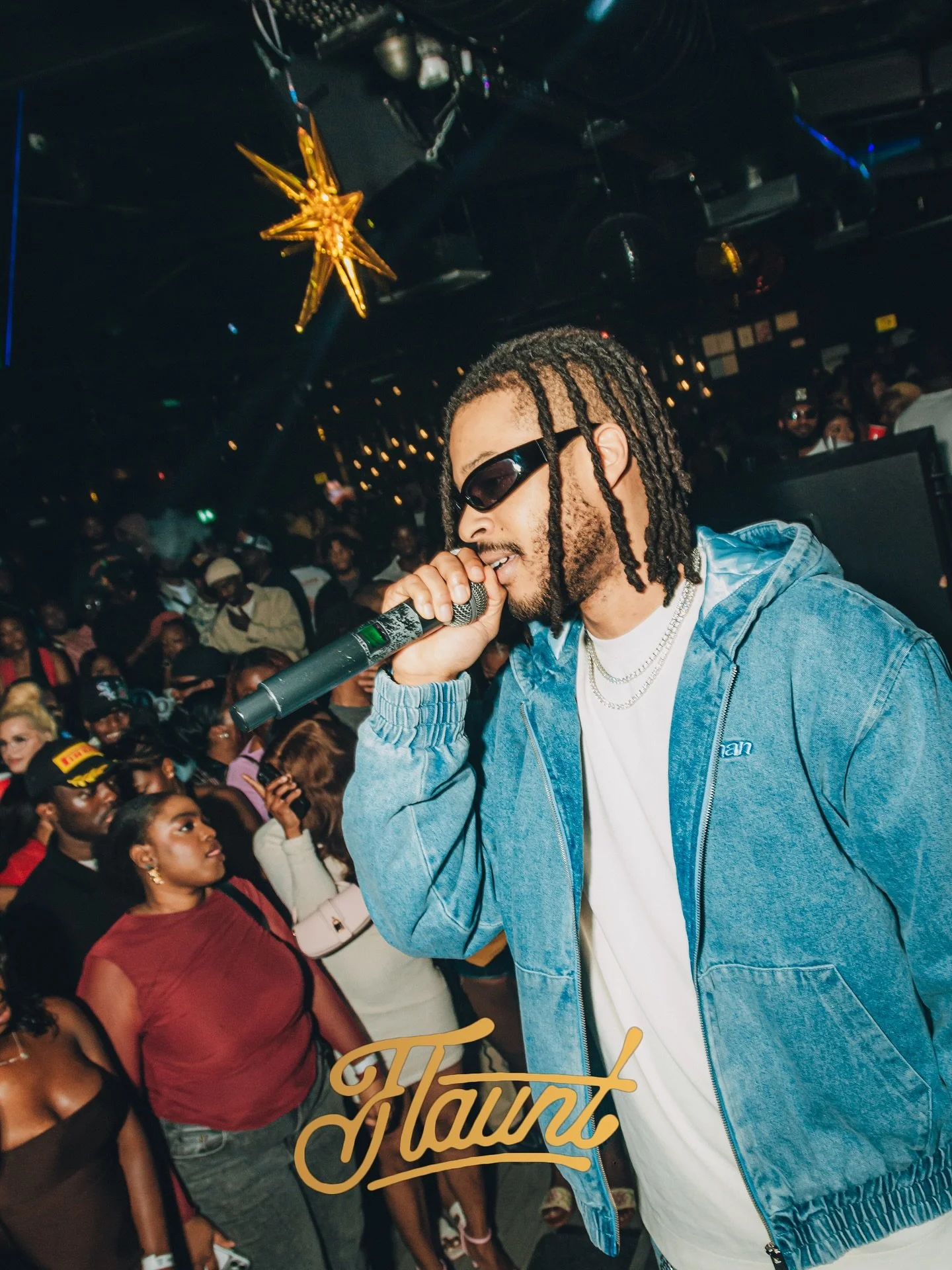 A male rapper with dreadlocks, sunglasses, and layered jewelry performs with a microphone at a crowded indoor concert, with a gold star hanging from the ceiling and the word 'Haut' in stylized script at the bottom.