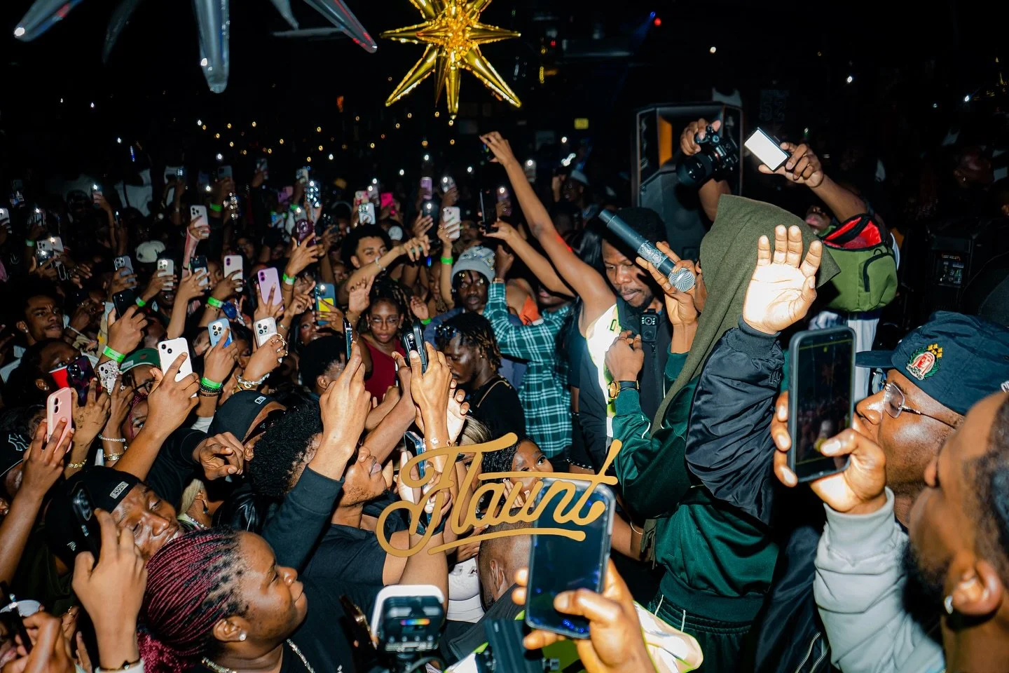 Mavo singing and surrounded by a crowded audience at a flaunt night, with many people holding up their phones to take pictures or record videos.